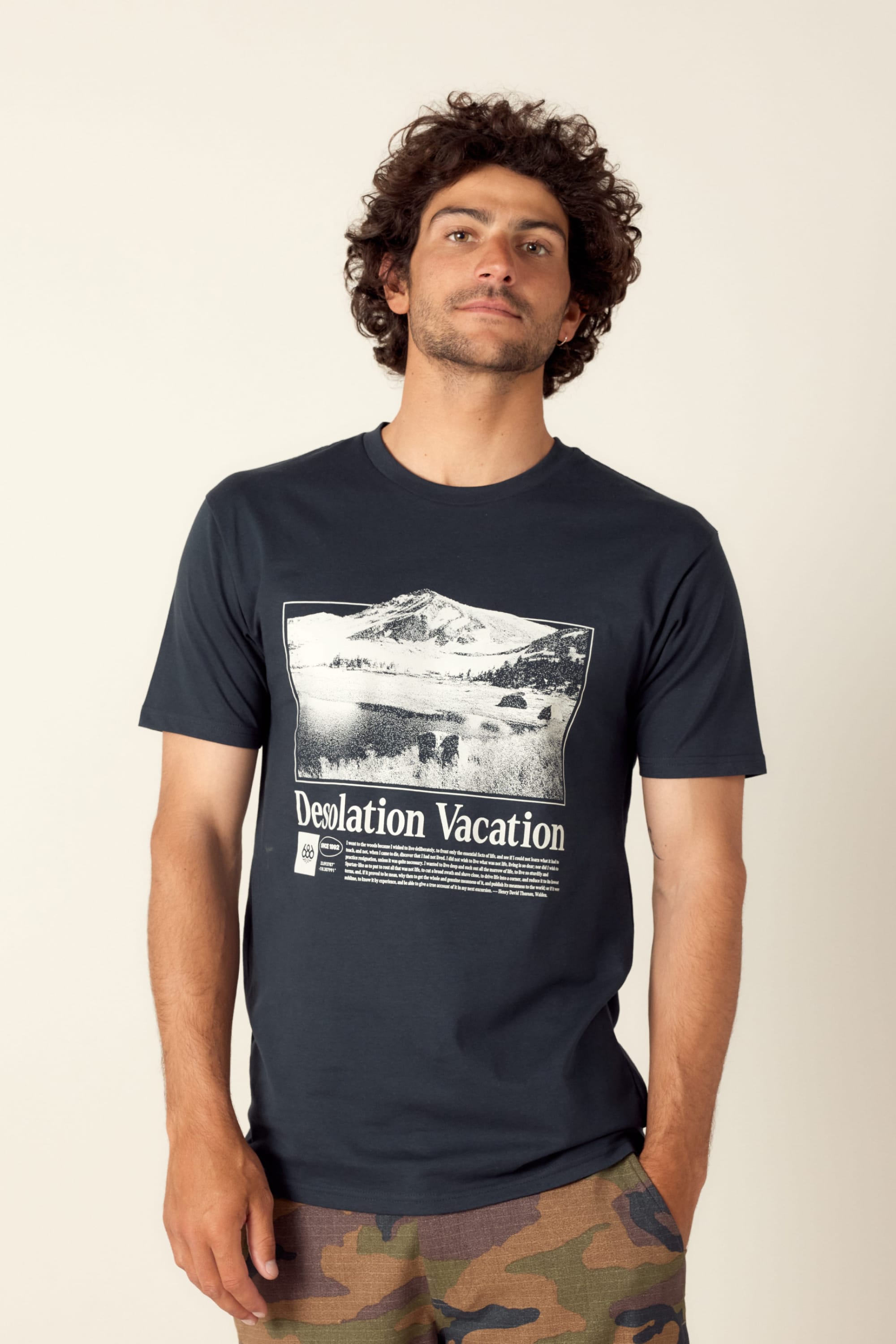 A man with curly hair wears the 686 Desolation Vacation Authentic S/S Tee by 686—featuring a mountain and lake graphic—paired with camouflage pants, standing against a plain light background.