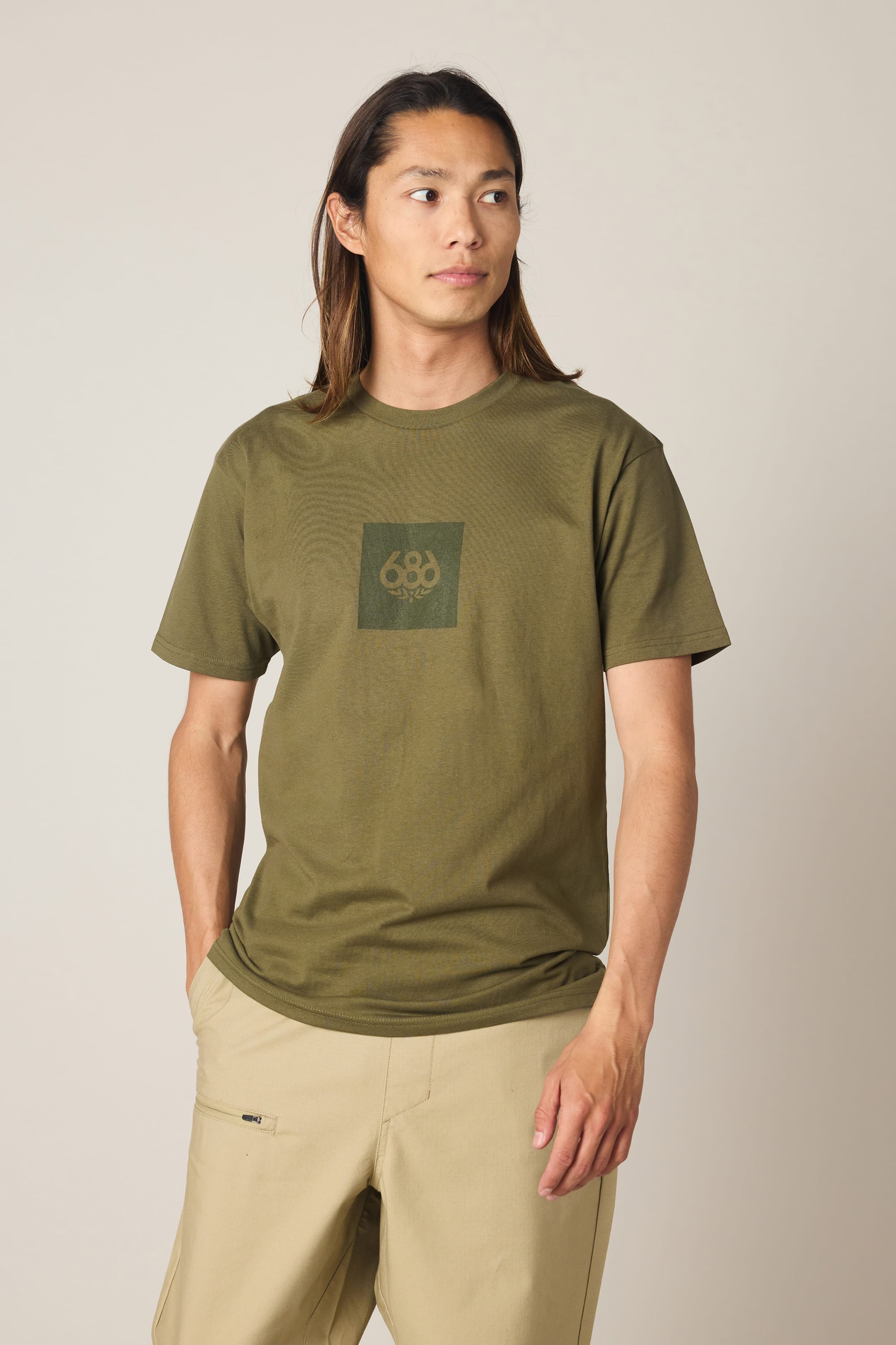 A person with long hair is wearing the 686 Knockout Authentic S/S Tee in olive green and beige pants, standing with hands in pockets and gazing slightly to the side against a plain background.