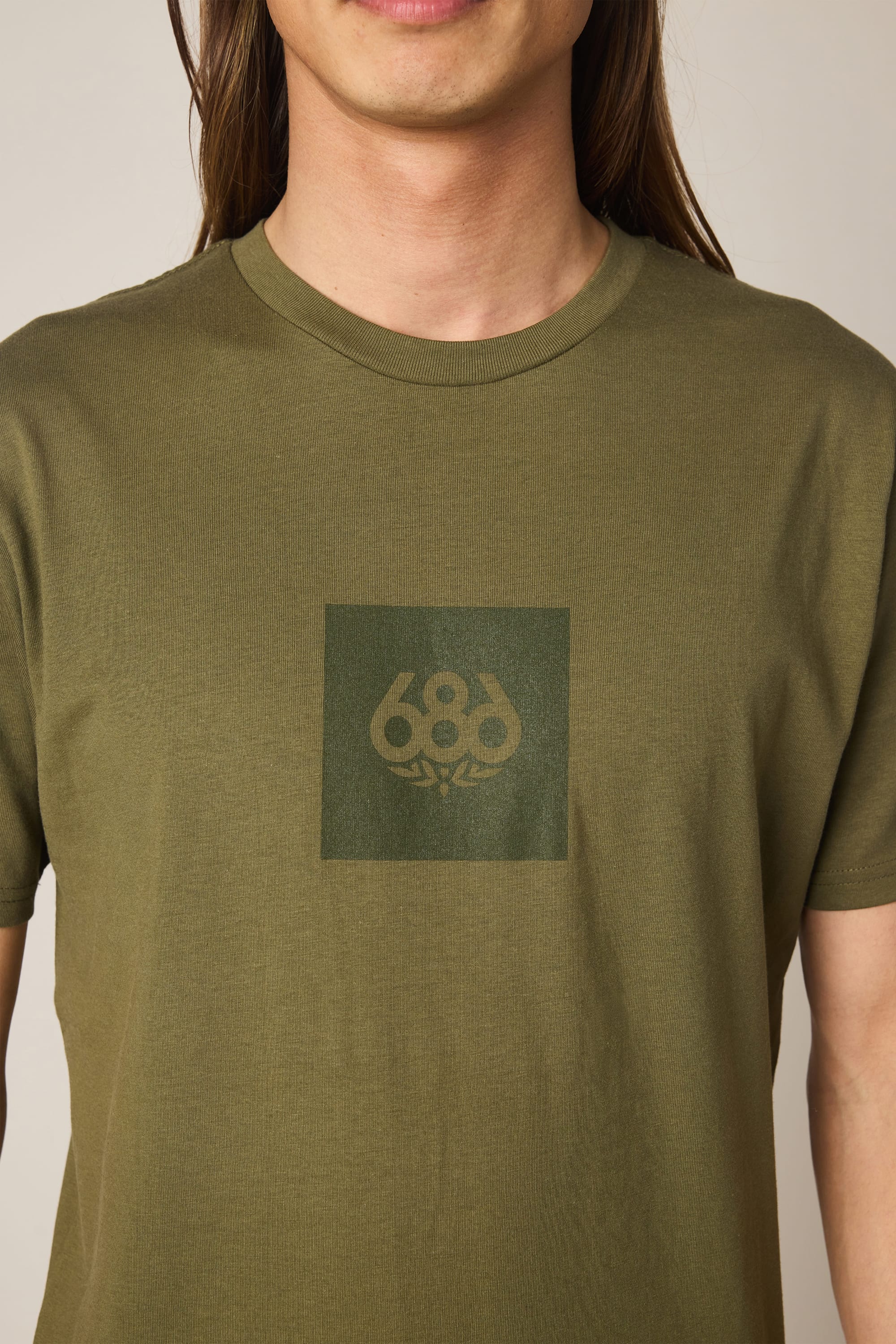 A person is shown wearing the 686 Knockout Authentic S/S Tee by 686, an olive green men’s cotton t-shirt with a dark green square and the 686 logo in the center. Only the upper torso and part of their face are visible.