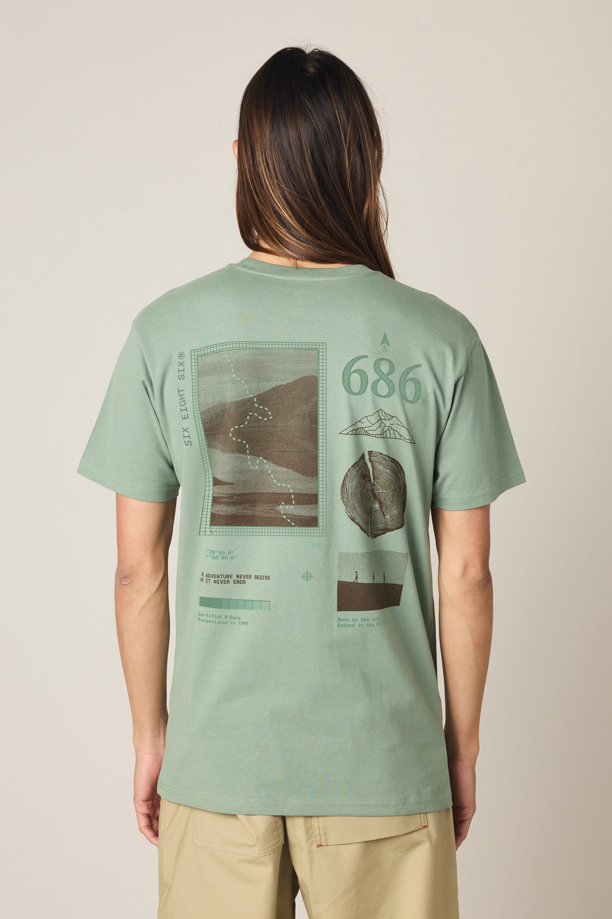 A person with long hair is wearing the 686 Path Authentic S/S Tee by 686, a light green t-shirt made from 100% cotton, featuring various graphic designs and text—including a map, mountain, and circular image—on the back; they are standing facing away from the camera.