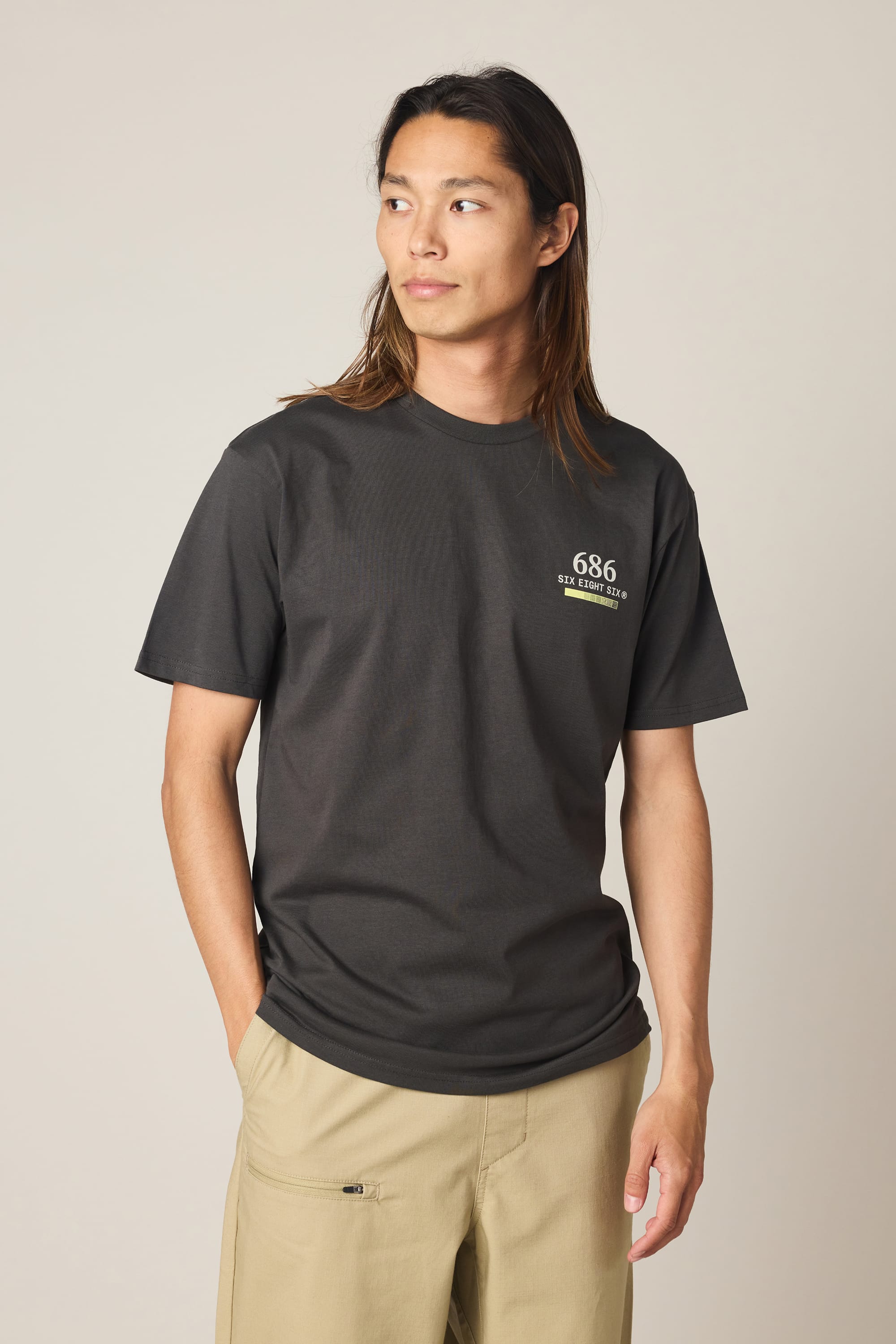 A person with long brown hair wears a black 686 Path Authentic S/S Tee from 686, made from 100% cotton, paired with light khaki pants. They stand with one hand in their pocket, looking to the side against a plain background.