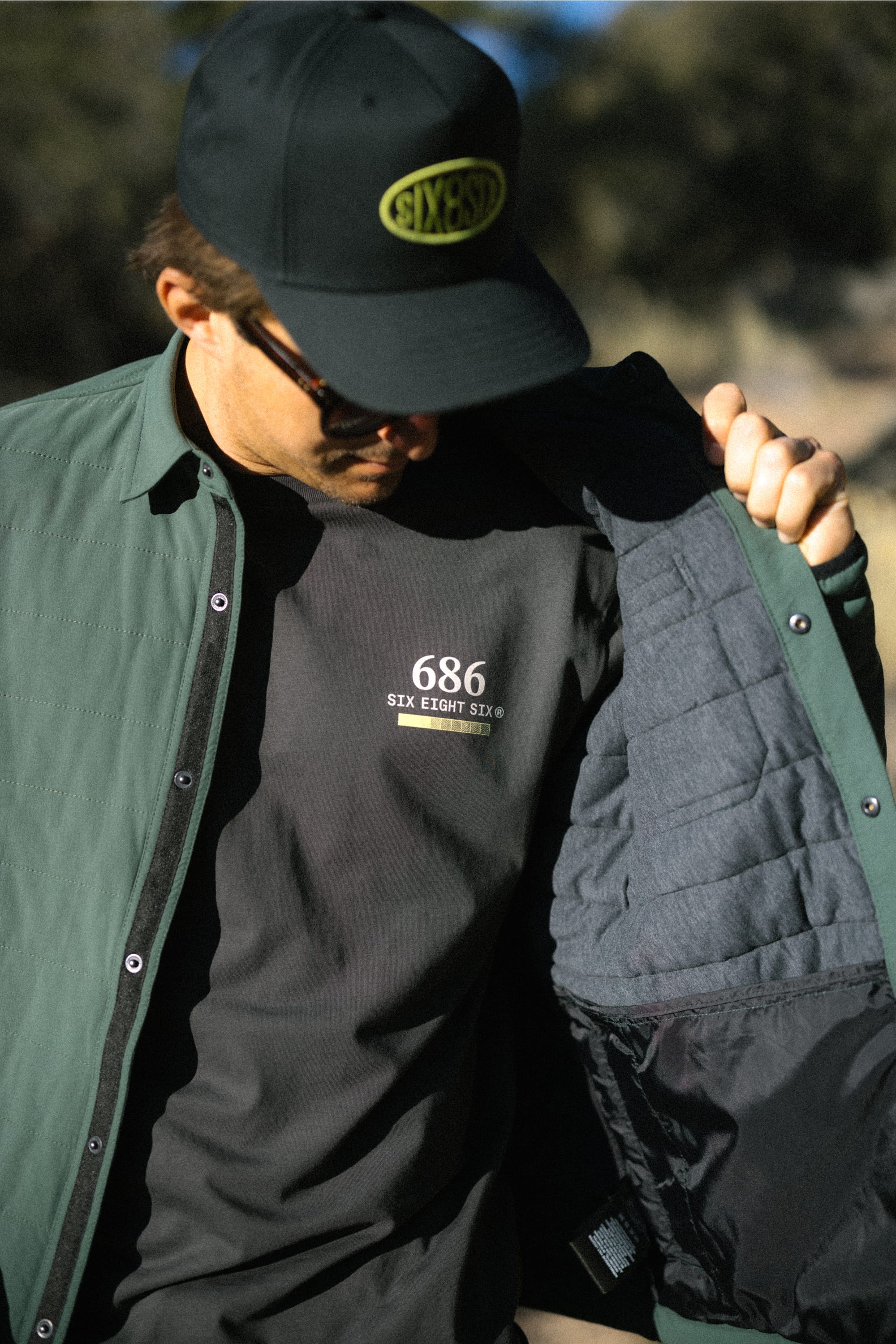 A person wearing a black cap and sunglasses opens their green jacket to reveal the 686 Path Authentic S/S Tee—a black men’s t-shirt by 686 made from 100% cotton for ultimate comfort, featuring “SIX EIGHT SIX” printed on the front.