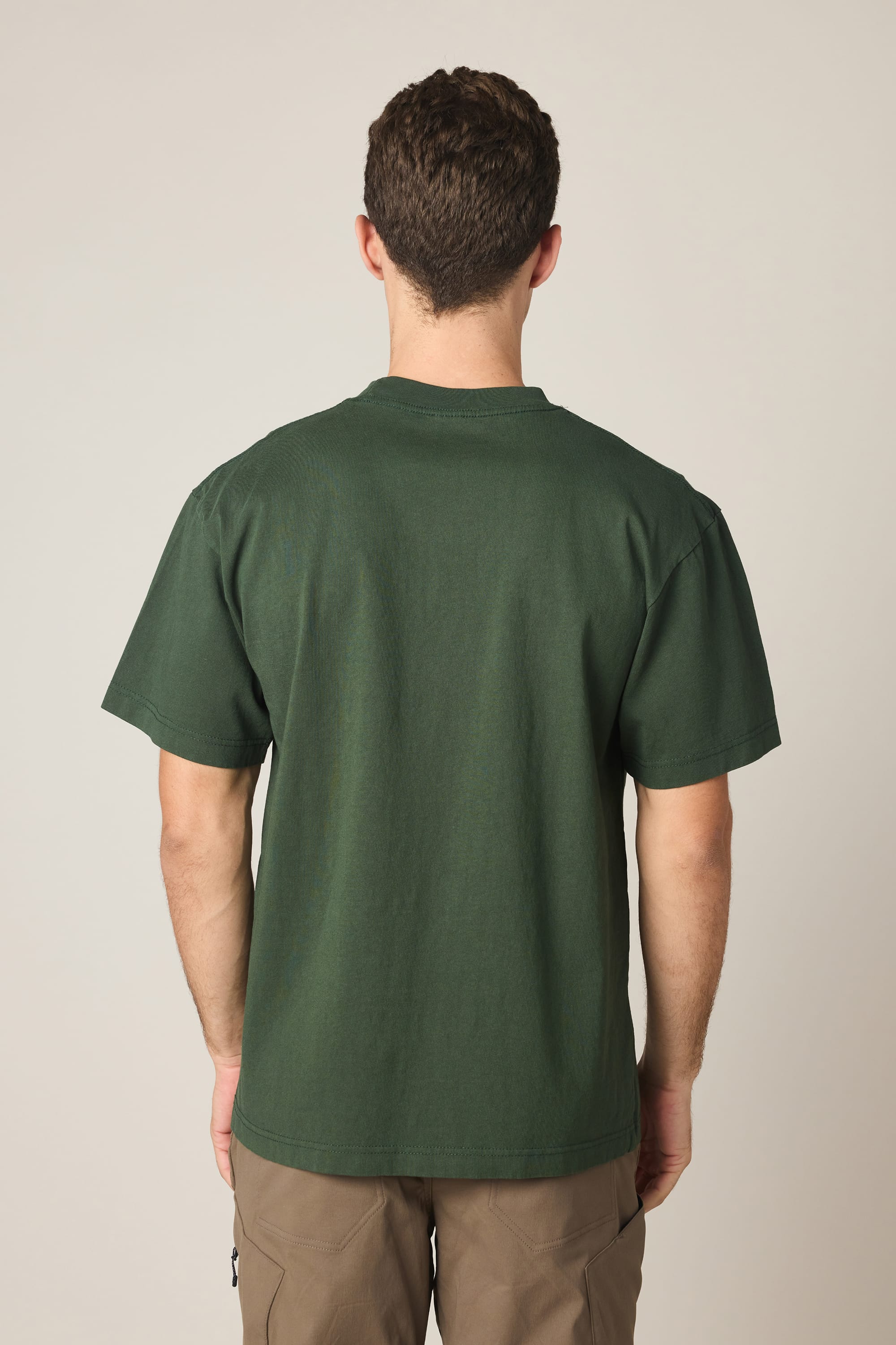 A man with short brown hair, wearing the 686 High Def Relaxed S/S Tee by 686 in dark green and brown pants, stands facing away from the camera against a plain light background.