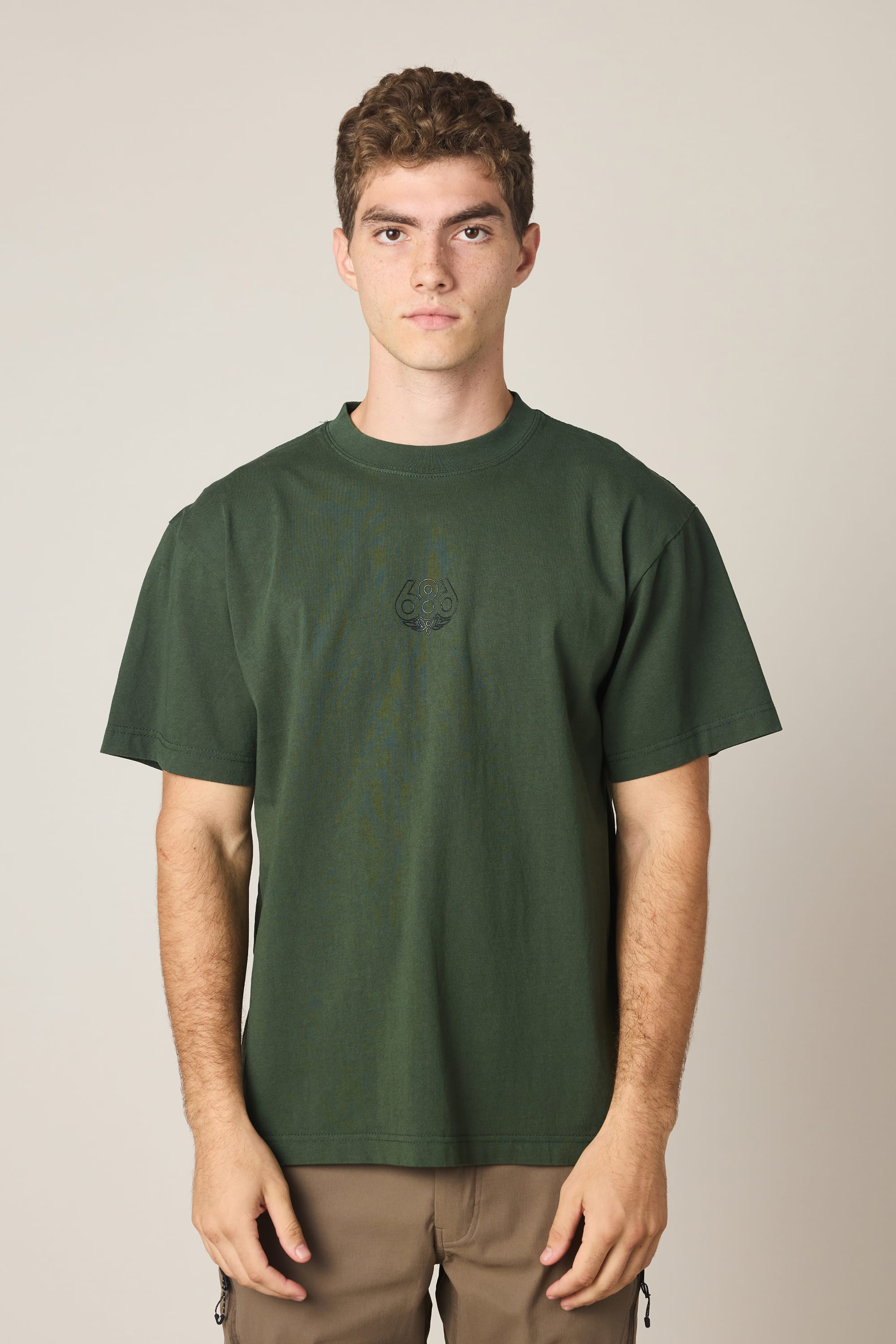 A young man with short curly hair wears the 686 High Def Relaxed S/S Tee by 686 in dark green and tan pants, standing against a plain light background, looking directly at the camera with a neutral expression.