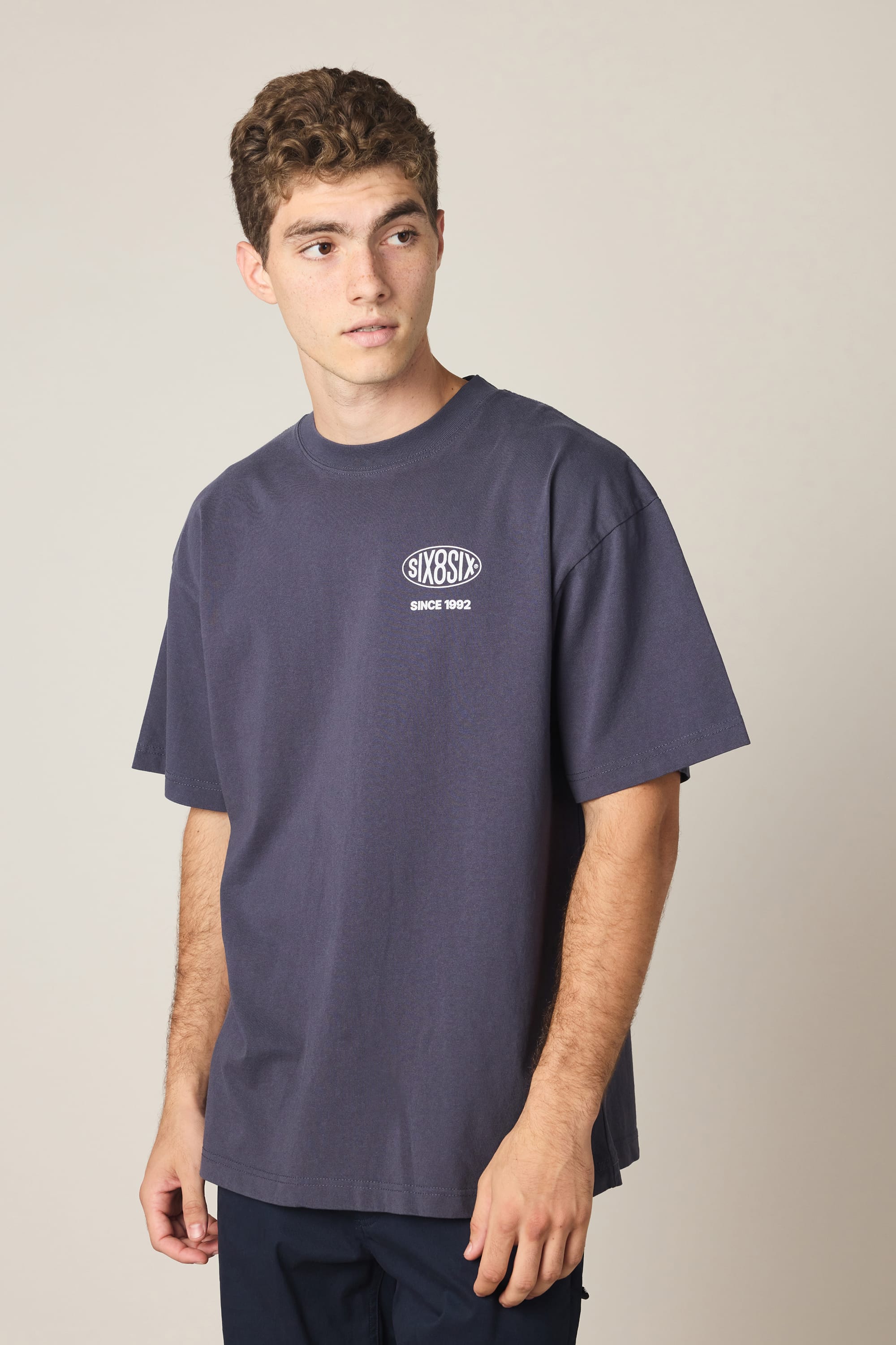 A young man with short curly hair wears the 686 92 Relaxed S/S Tee by 686—a loose-fitting, dark gray men’s T-shirt made from 100% cotton, featuring a small white “SIXOZ SINCE 1992” logo and text on the left chest. He pairs it with navy pants and looks off to his left.