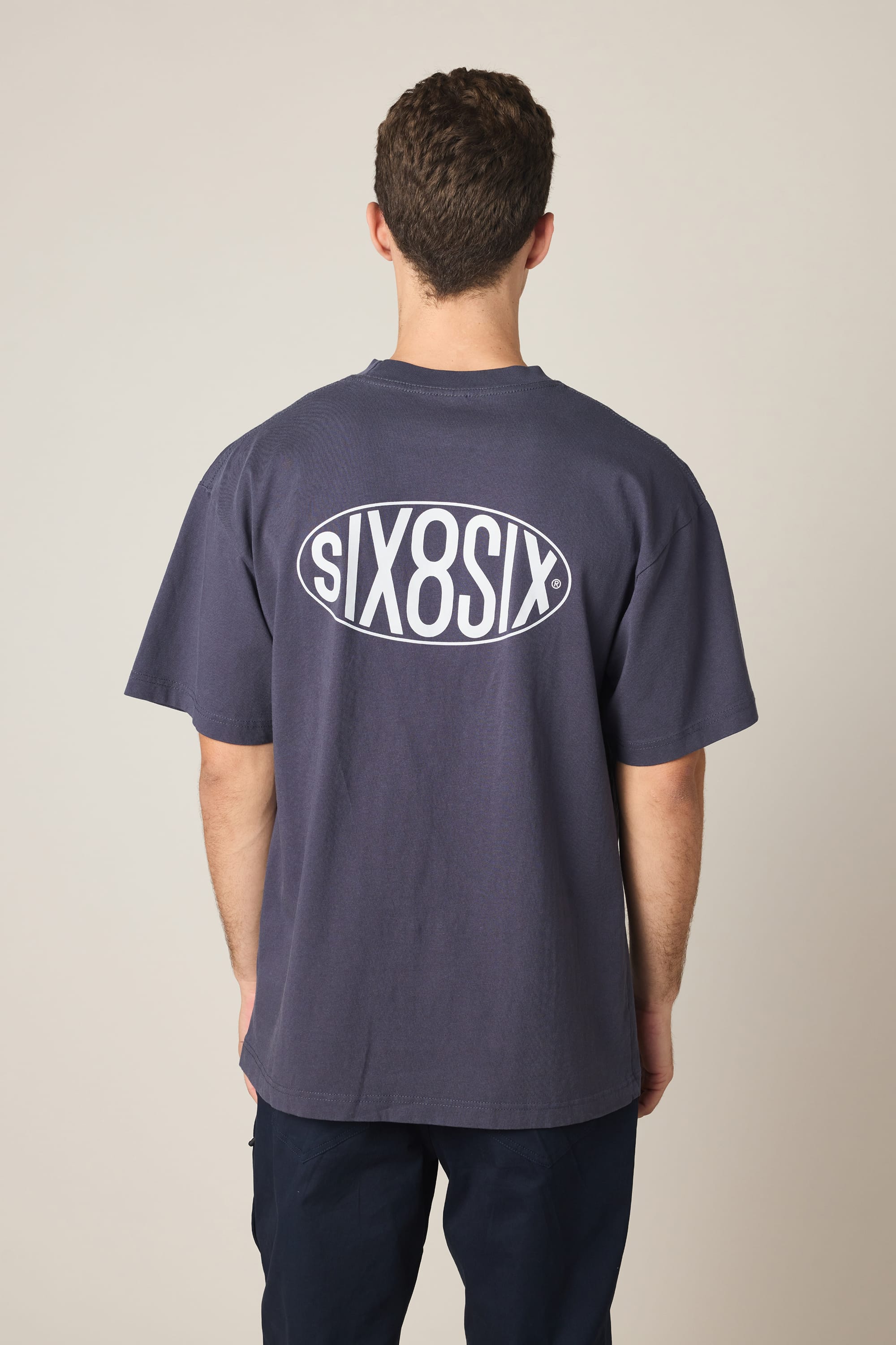 A person stands facing away from the camera, wearing the 686 92 Relaxed S/S Tee—a men's dark blue t-shirt by 686 featuring a large white SIX8SIX oval logo on the back. The 100% cotton fabric stands out against the plain, light-colored background.