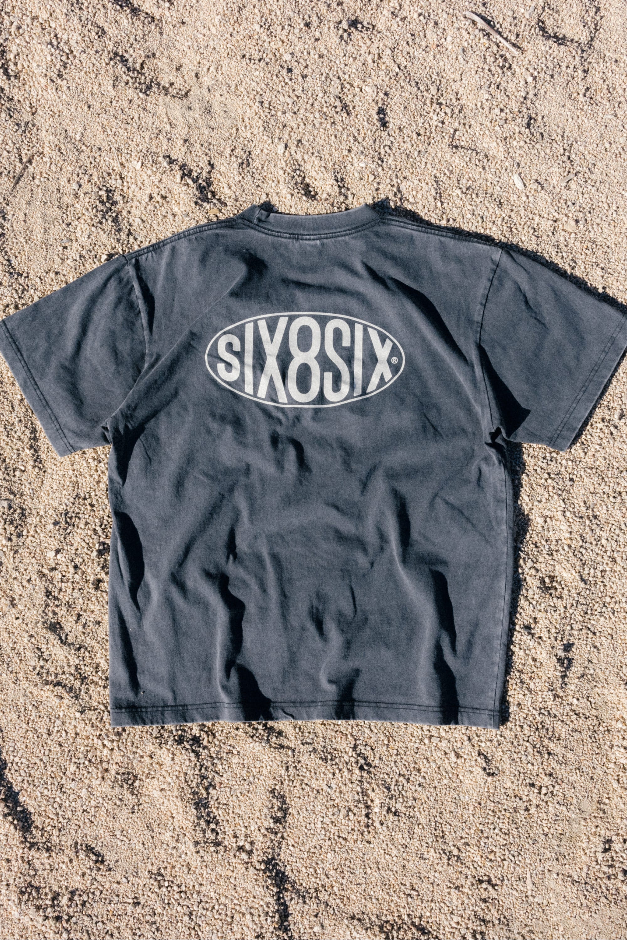 A black 686 92 Relaxed S/S Tee by 686 lies on sandy ground, showcasing a large oval-shaped logo with bold white SIX8SIX lettering on the back. Made from 100% cotton, this t-shirt delivers comfort and style for everyday wear.