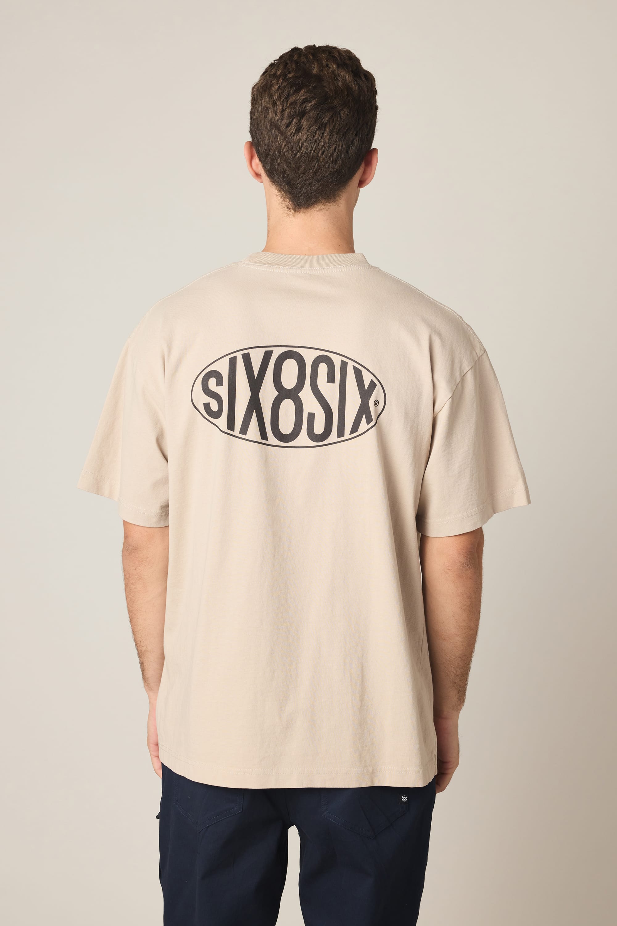 A person stands with their back to the camera, wearing the 686 92 Relaxed S/S Tee in beige. This men's t-shirt by 686 features "SIX8SIX" in bold black letters inside an oval on the back and is made from soft 100% cotton for comfortable, all-day wear.