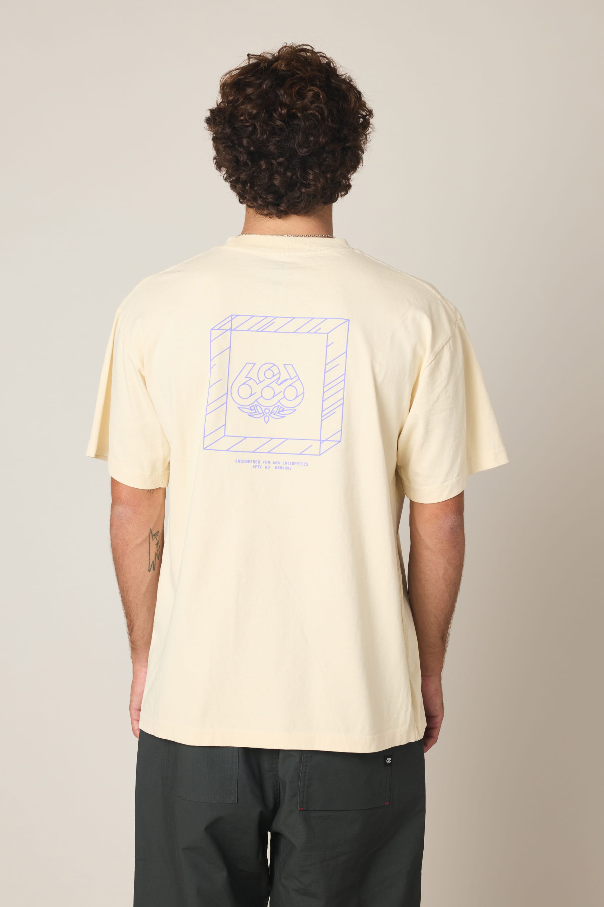 A person with curly hair is seen from behind wearing the 686 Engineered Relaxed SS Tee‚Äîa cream men's 100% cotton shirt with a purple geometric graphic and ‚Äú686‚Äù on the back‚Äîpaired with dark pants against a plain, light background.