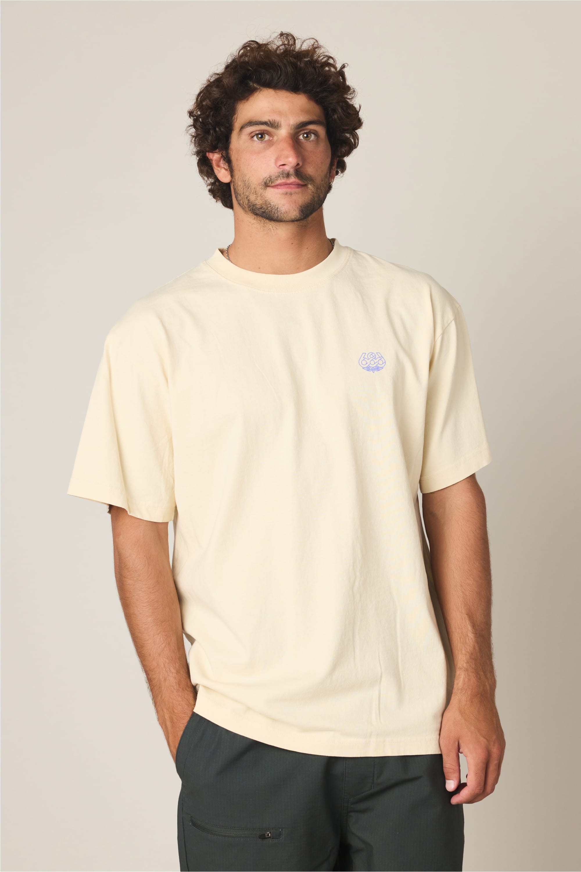 A man with curly brown hair and a beard stands against a plain background, wearing the 686 Engineered Relaxed SS Tee by 686‚Äîa loose, light yellow 100% cotton men's t-shirt with a small logo on the chest‚Äîand dark pants. He looks slightly off to the side.