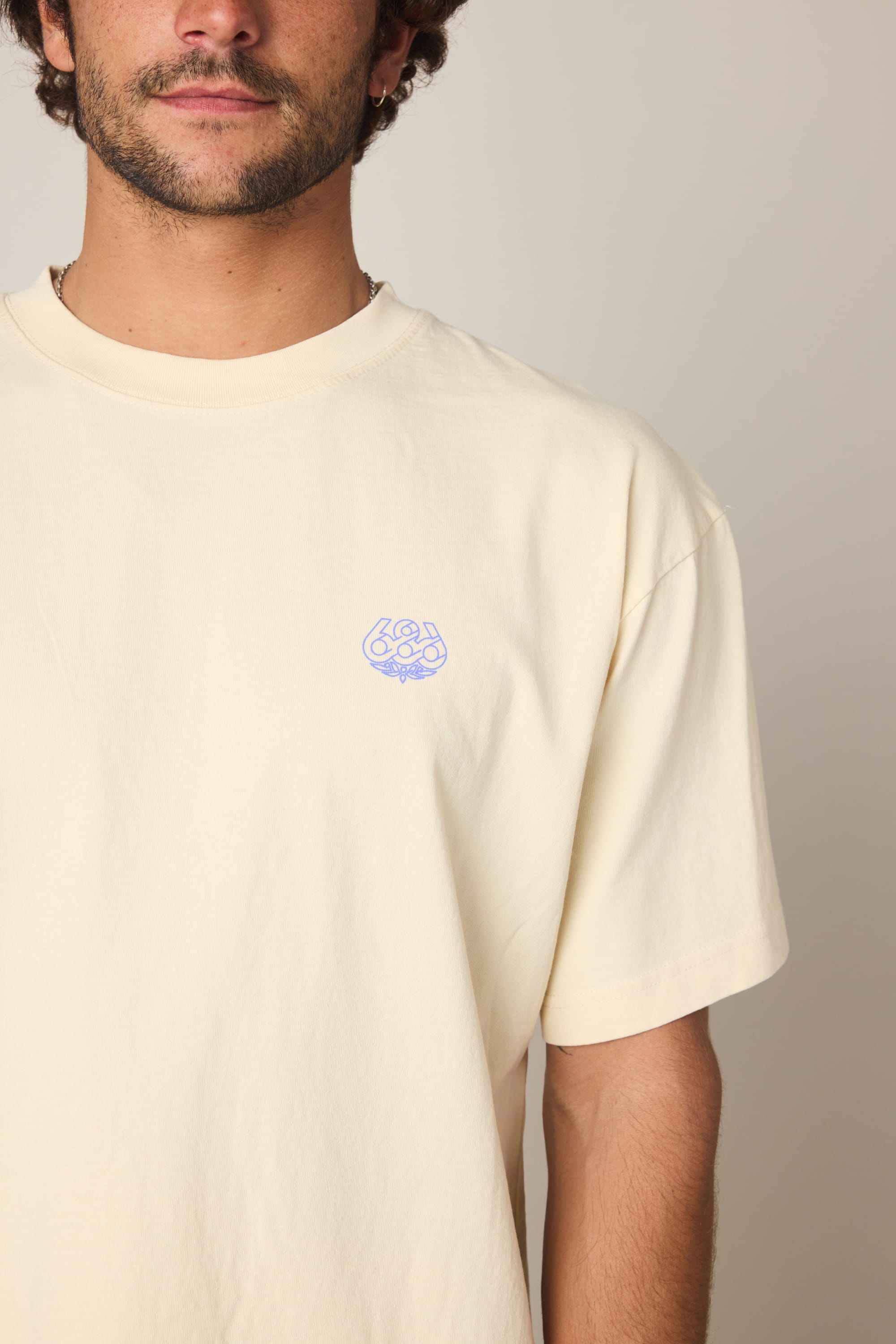 A person wears the 686 Engineered Relaxed SS Tee, a light beige men's short-sleeve shirt featuring a small purple logo on the upper left chest. Made by 686 from 100% cotton, it looks comfortable and soft. The person's face is partly out of frame; background is plain.