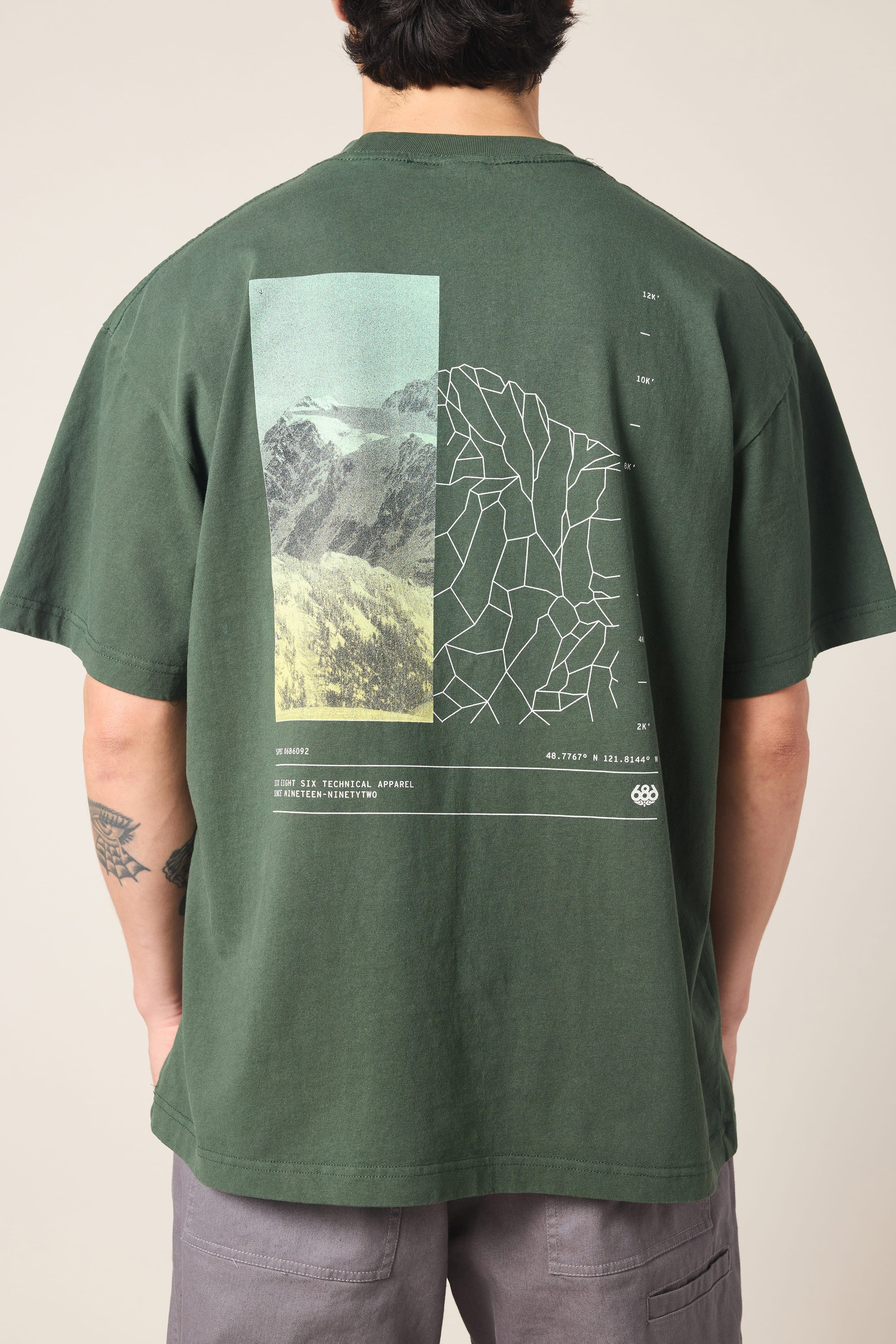 A person stands with their back to the camera, wearing the 686 Peaks Relaxed SS Tee by 686‚Äîa men's dark green 100% cotton t-shirt featuring a mountain photo, geometric line design, and small white text graphic on the back.