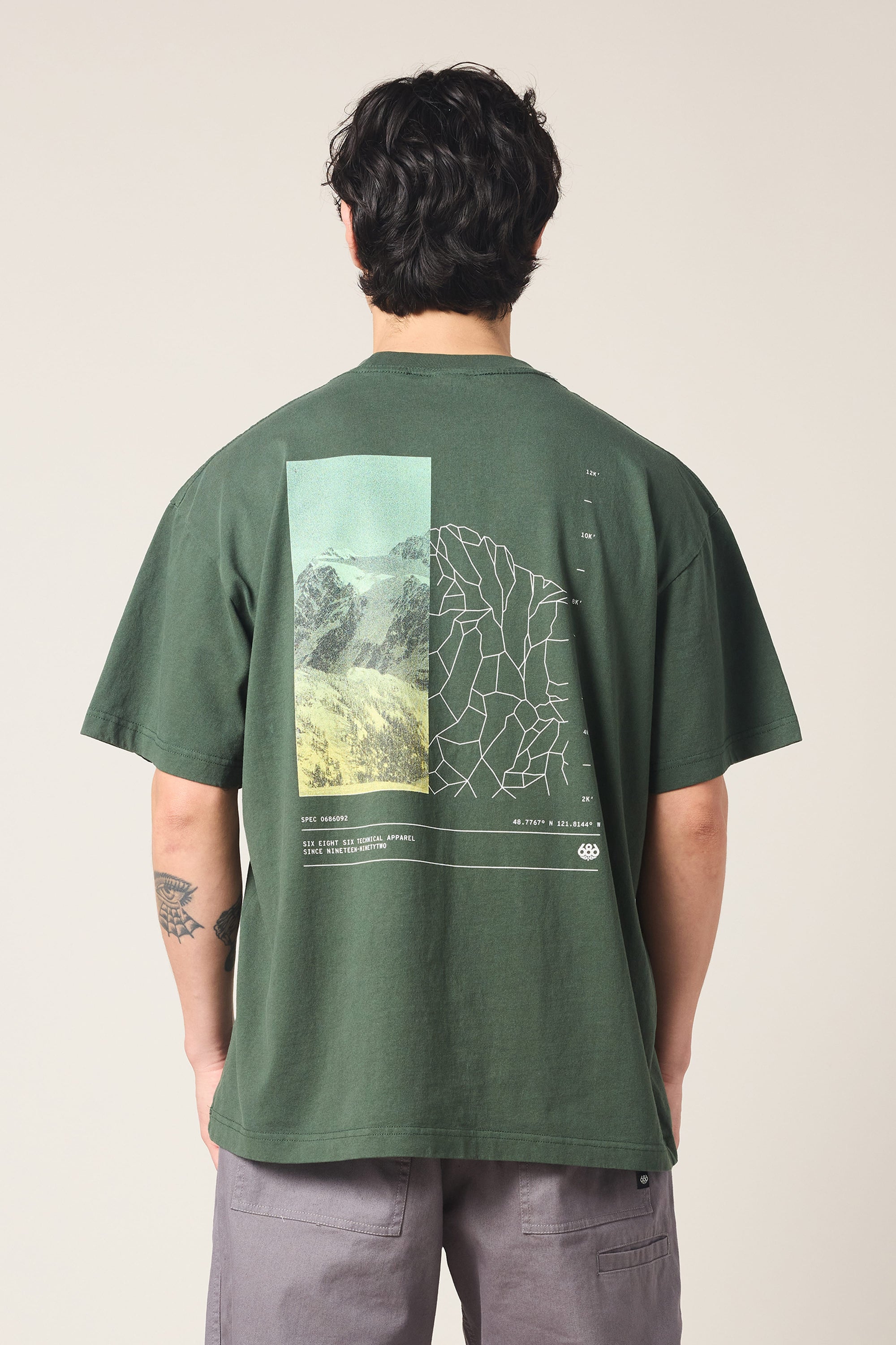 A person with dark hair and a left arm tattoo stands facing away, wearing the 686 Peaks Relaxed SS Tee by 686‚Äîa dark green 100% cotton men's t-shirt featuring a mountain and abstract line graphic on the back.