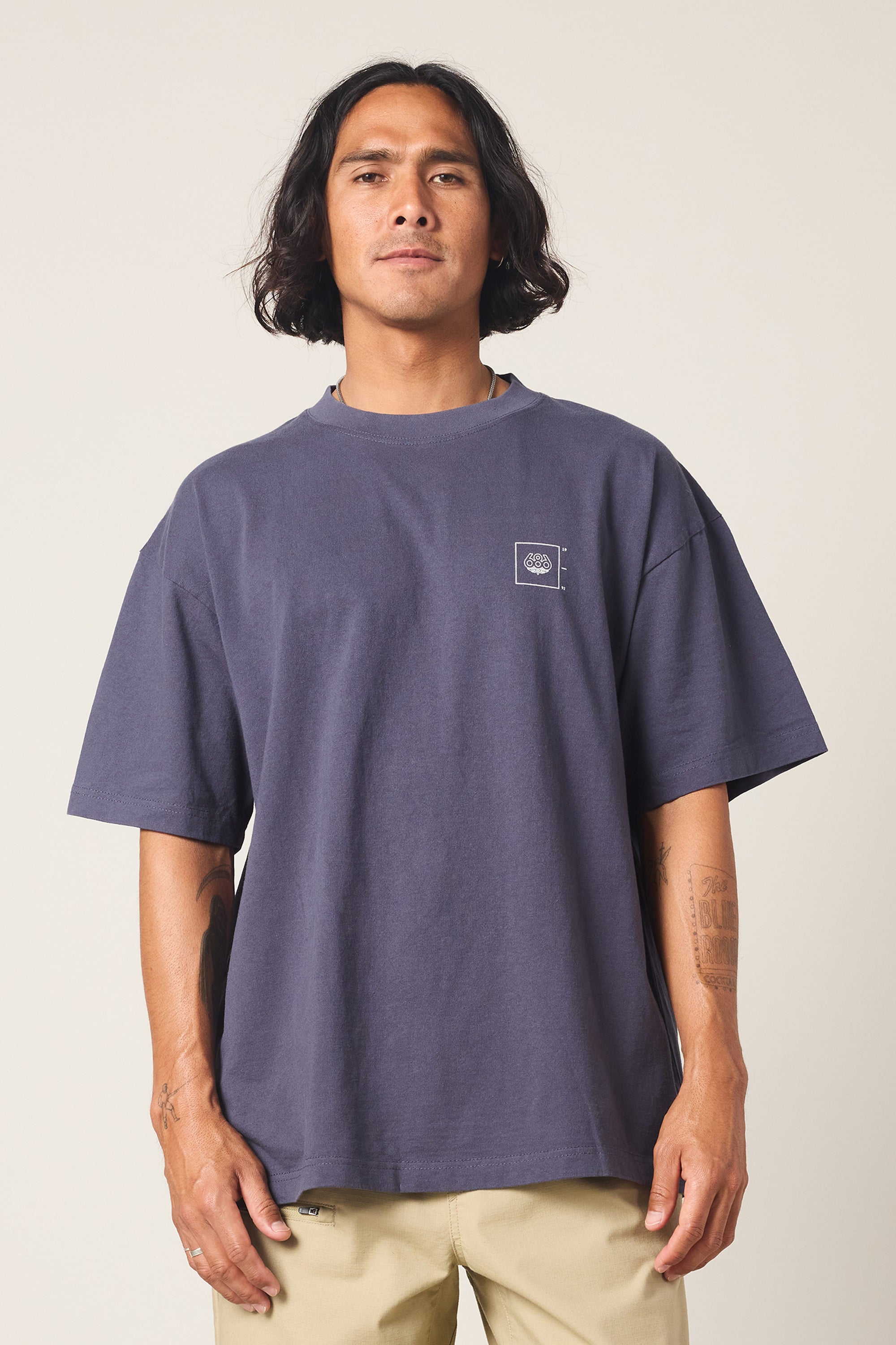 A person with medium-length dark hair wears the 686 Peaks Relaxed SS Tee, a loose-fitting men's cotton navy shirt with a small white logo on the chest, paired with light beige pants, standing against a plain light background.