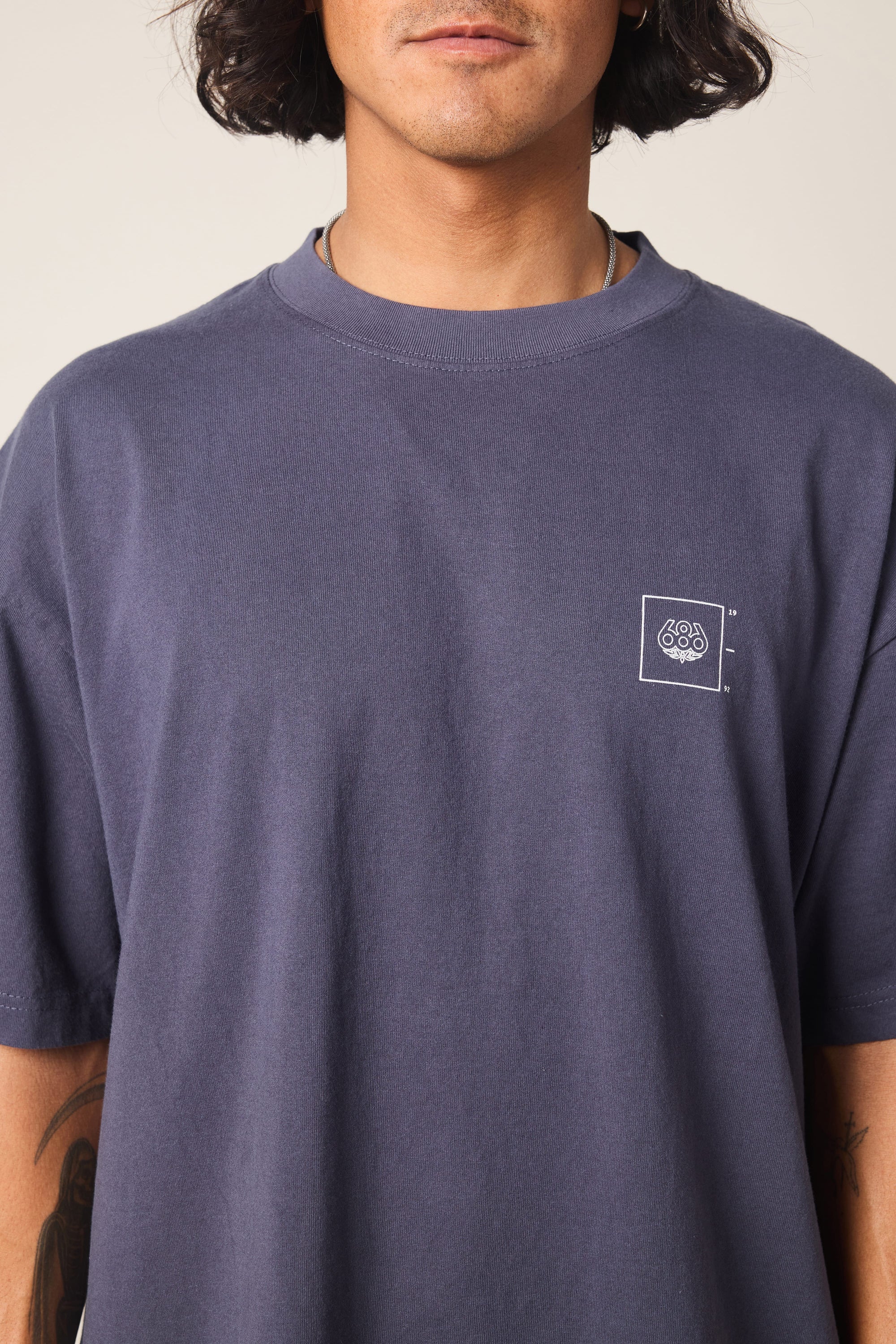 A person with medium-length dark hair wears the 686 Peaks Relaxed SS Tee by 686‚Äîa loose-fitting, faded purple men's T-shirt made from 100% cotton with a small white geometric logo on the left chest. Photo cropped nose to waist.