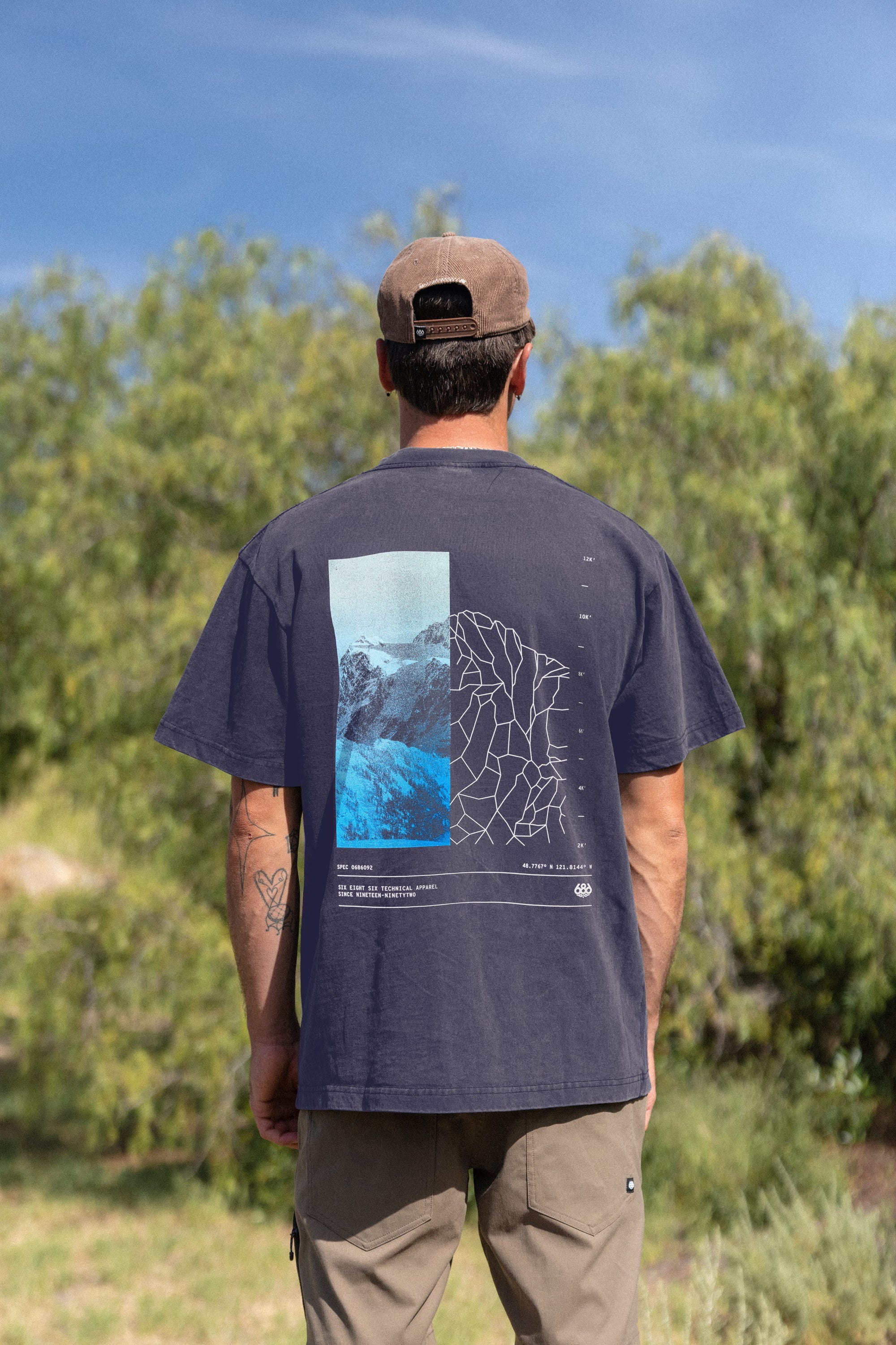 A man stands outdoors among green trees under a blue sky, wearing the 686 Peaks Relaxed SS Tee by 686‚Äîa black, 100% cotton tee with abstract graphics and a mountain photo on the back‚Äîpaired with a brown cap and light khaki pants.