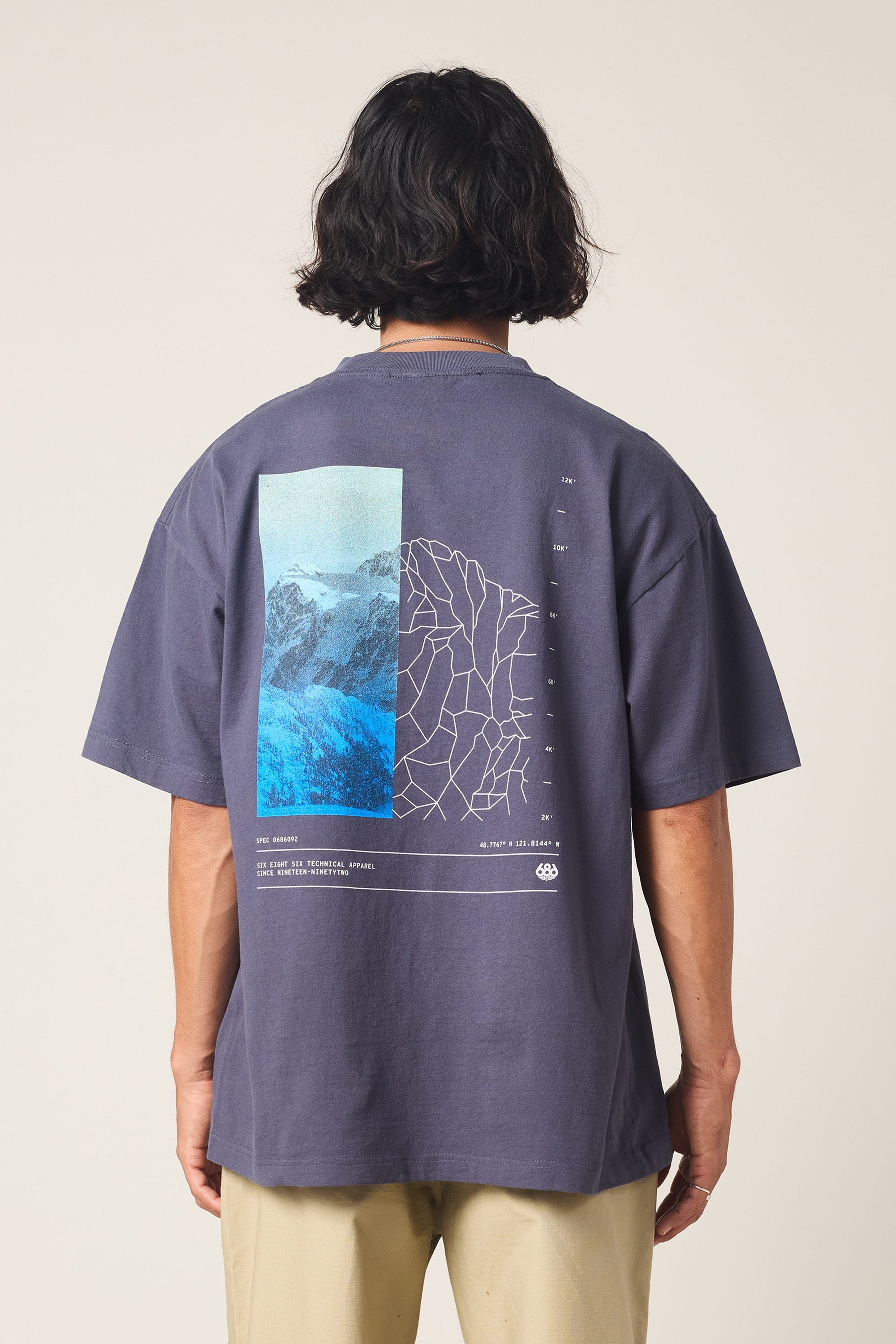 A person with medium-length dark hair, seen from the back, wears the 686 Peaks Relaxed SS Tee by 686‚Äîa dark gray 100% cotton men's shirt featuring a blue mountain and white contour map graphic‚Äîwith beige pants.