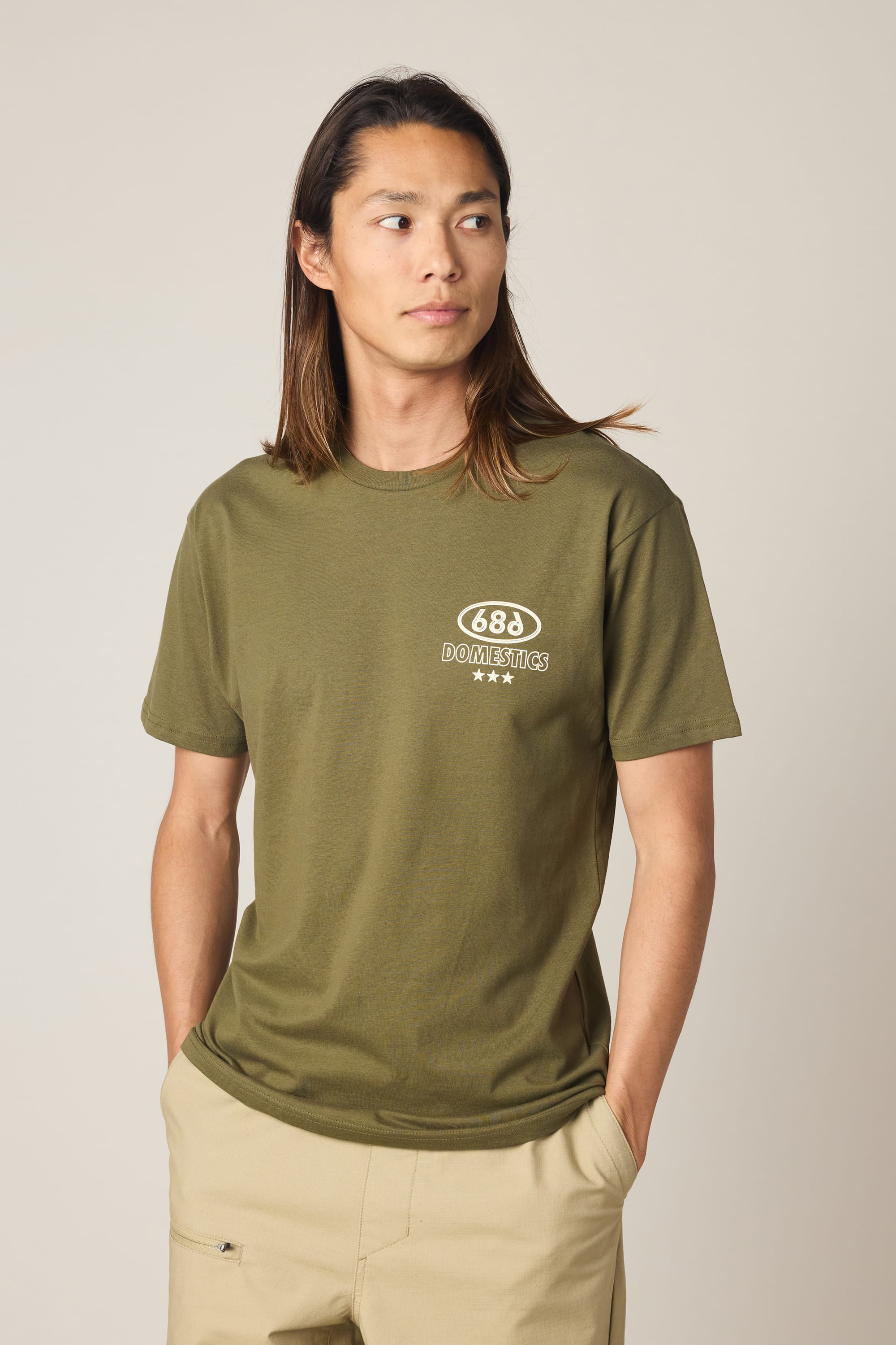 A person with long brown hair wears the 686 Men's Domestics Authentic SS Tee in olive green with a small white 688 logo, paired with beige pants, standing against a plain light background and looking to the side with hands in pockets.
