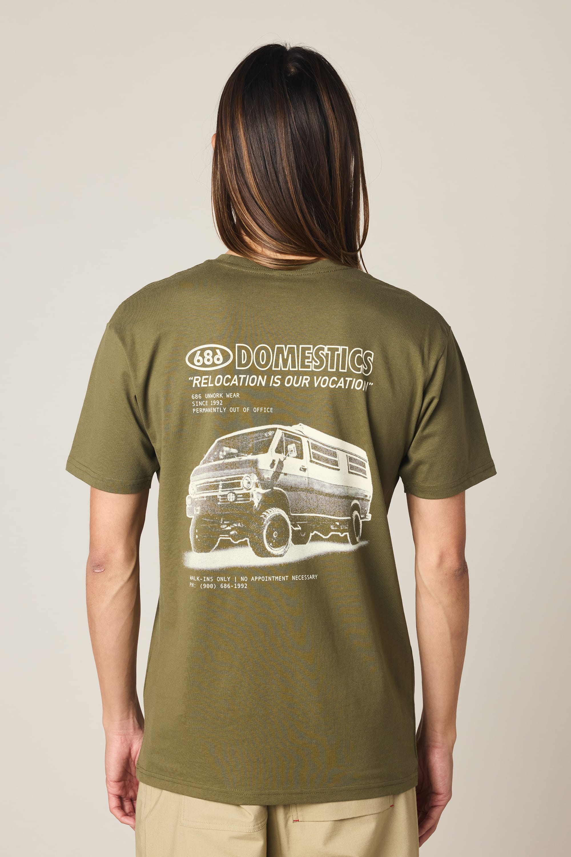 Person with long hair, seen from the back, wears a 686 Men's Domestics Authentic SS Tee by 686—an olive green 100% cotton shirt with a white van graphic and text: “688 Domestics. Relocation is our vocation.” plus small details.