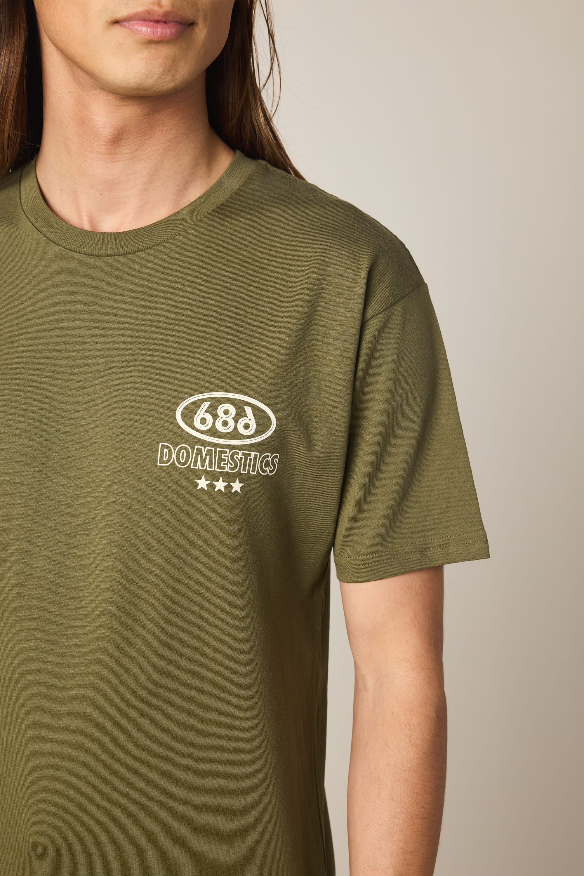 A man wears the 686 Men's Domestics Authentic SS Tee in olive green, featuring "68d DOMESTICS" and three white stars on the left chest. Made from 100% cotton, only his upper torso and part of his face are shown.