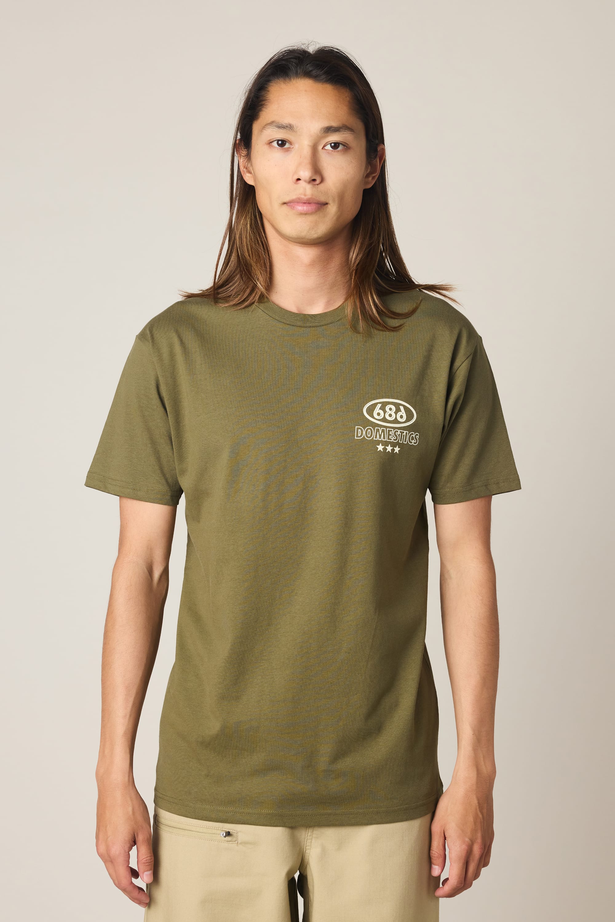 A person with long brown hair stands facing forward, wearing the 686 Men's Domestics Authentic SS Tee in olive green with a small white 686 logo on the upper left chest and light beige pants, set against a plain background.