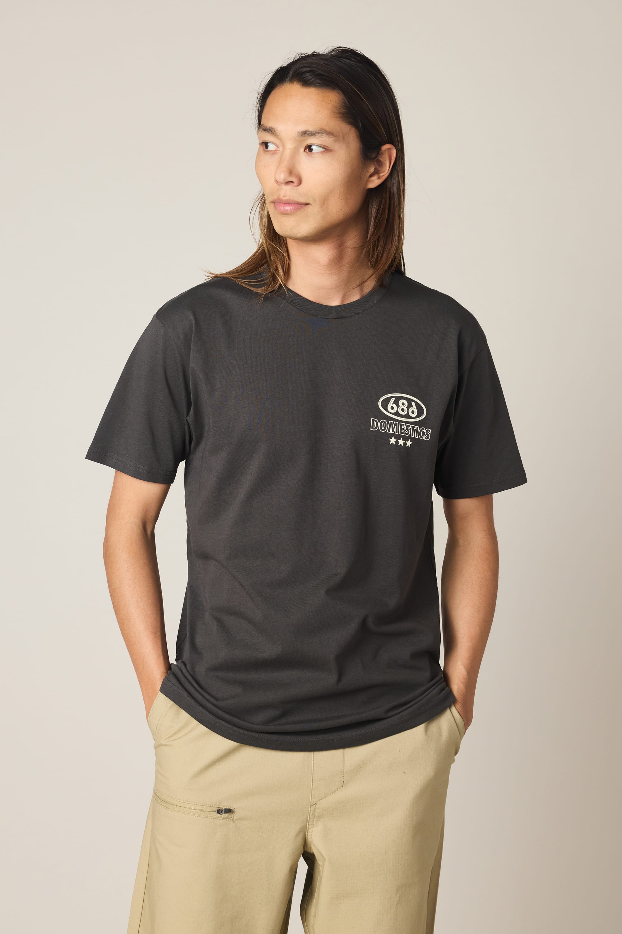 A person with long brown hair wears the 686 Men's Domestics Authentic SS Tee by 686, a dark gray 100% cotton shirt with a small white left-chest graphic, paired with khaki pants, standing hands in pockets against a plain background.