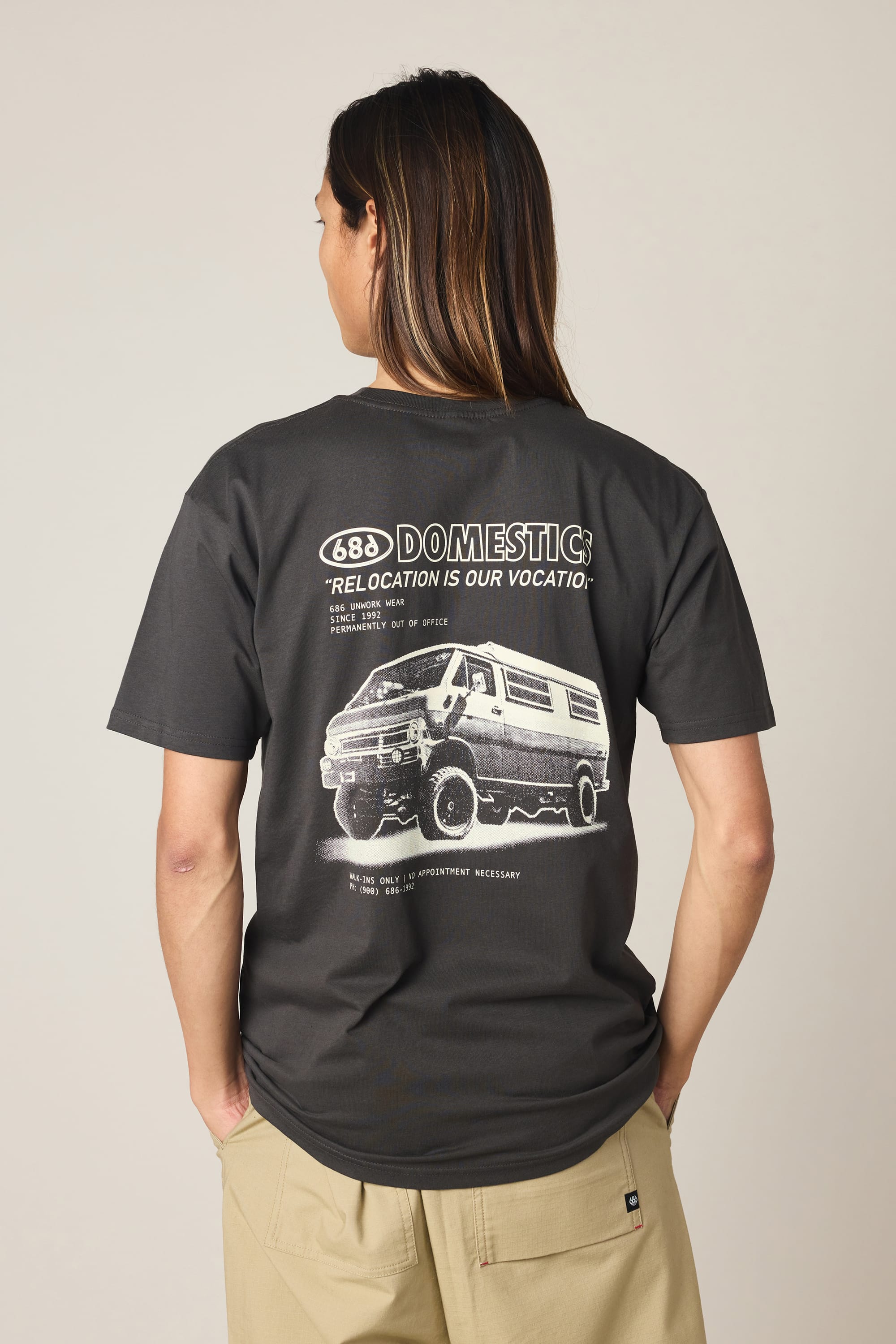 A person with long hair, seen from behind, wears the 686 Men's Domestics Authentic SS Tee by 686, a black 100% cotton shirt with a van graphic and “DOMESTIC: Relocation is our vocation” text on the back, paired with beige pants.