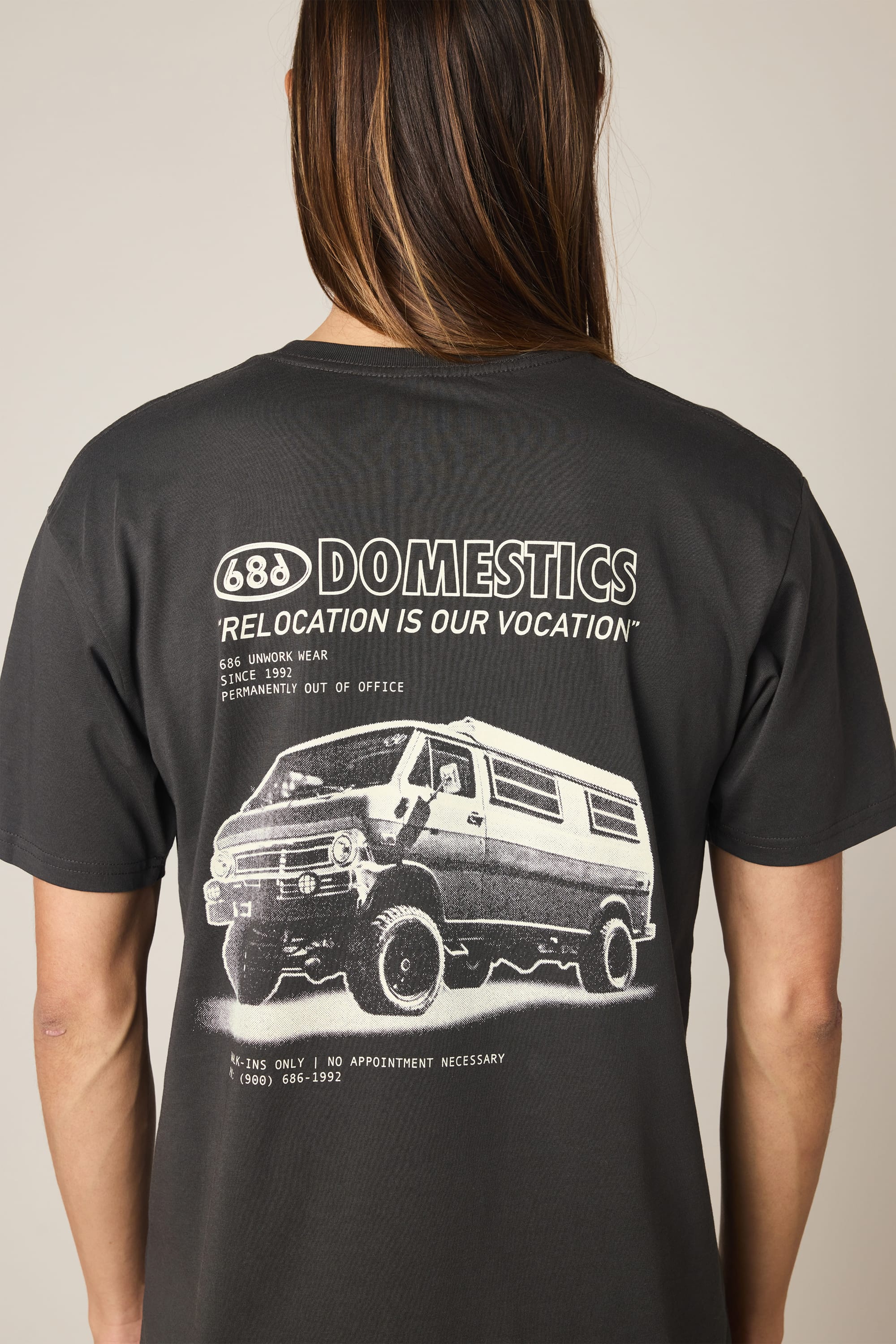 A person wearing the 686 Men's Domestics Authentic SS Tee—a black 100% cotton t-shirt by 686—featuring white graphic text reading "DOMESTICS RELOCATION IS OUR VOCATION" and an image of a van on the back, along with additional small text above and below the illustration.