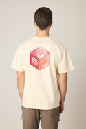 A person wears the 686 Men's Unit Relaxed SS Tee by 686, a cream 100% cotton t-shirt with a large red and pink die graphic showing numbers 6, 8, and 0 on the back, standing against a plain light background.