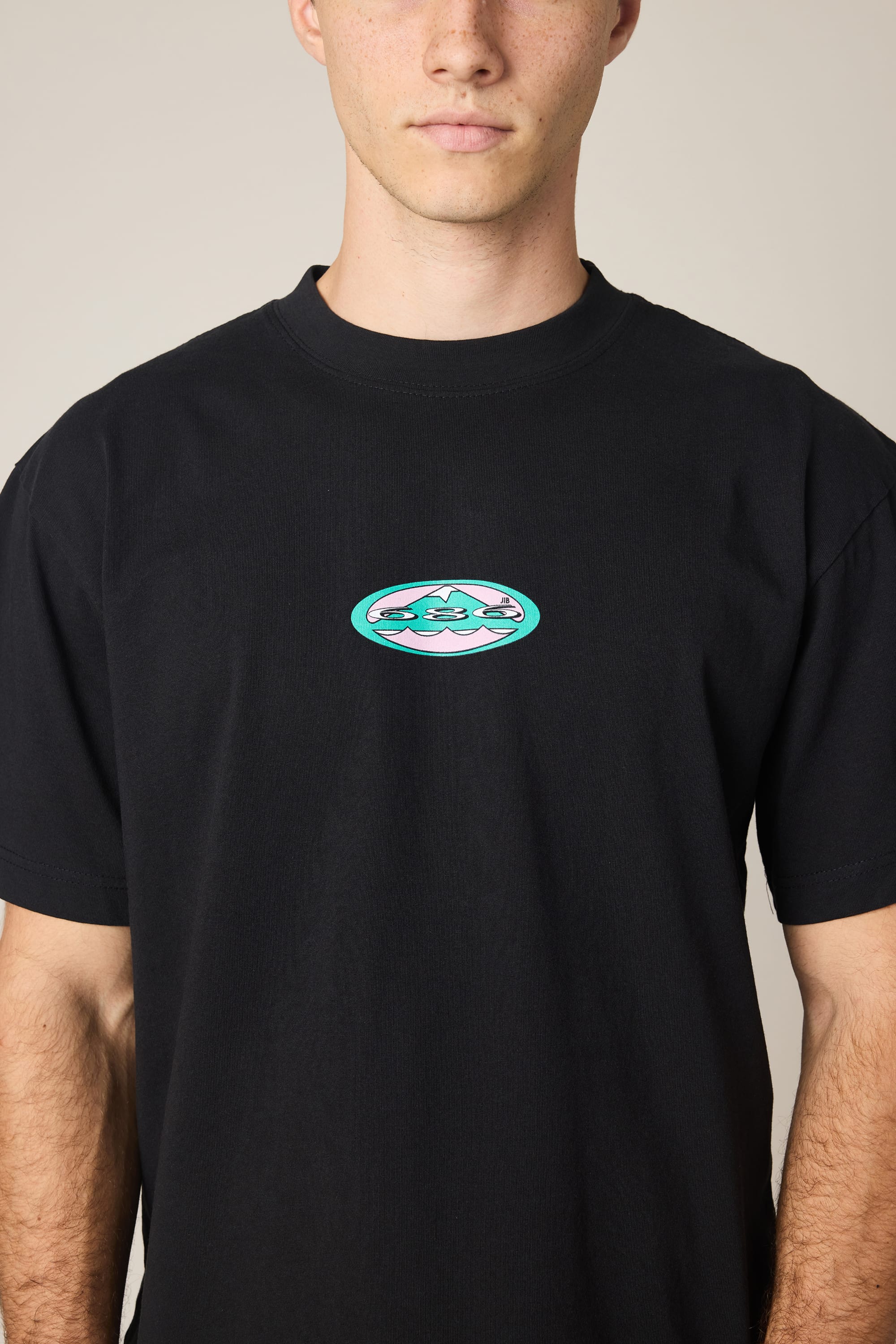 A person wearing the 686 Men's Jib Relaxed SS Tee, a black 100% cotton shirt by 686 featuring a small, colorful three-face graphic inside an oval on the chest; photo cropped from shoulders to waist.