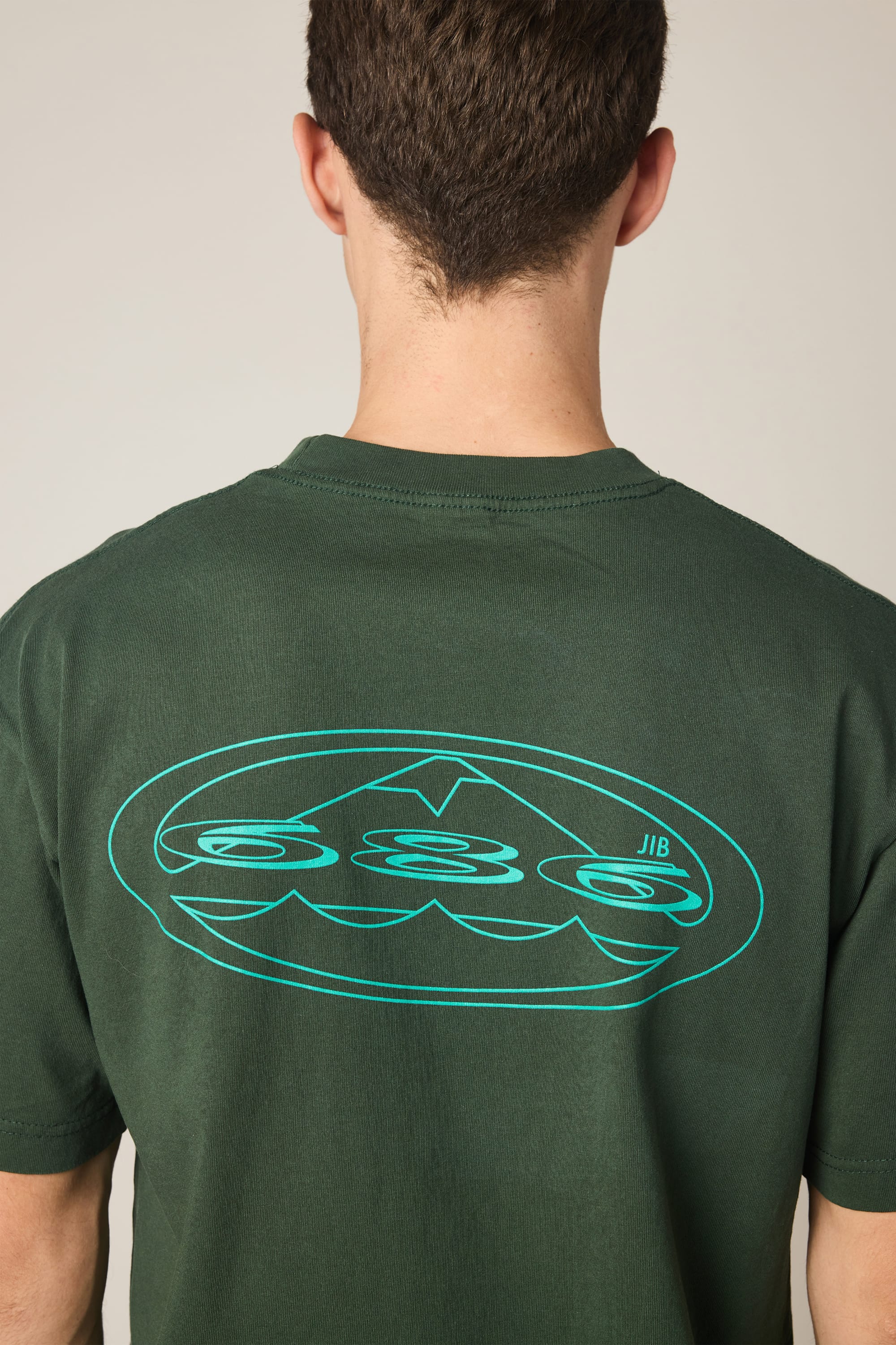A person wearing the 686 Men's Jib Relaxed SS Tee in dark green, viewed from behind. This 100% cotton t-shirt by 686 features a large, stylized blue graphic with abstract shapes and lines on the upper back.