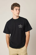 A young man with short curly hair wears the 686 Men's Unwork Spec Relaxed SS Tee, a black 100% cotton t-shirt featuring a small white 686 logo and text on the left chest. He stands with hands in his beige pants pockets, looking to the side.