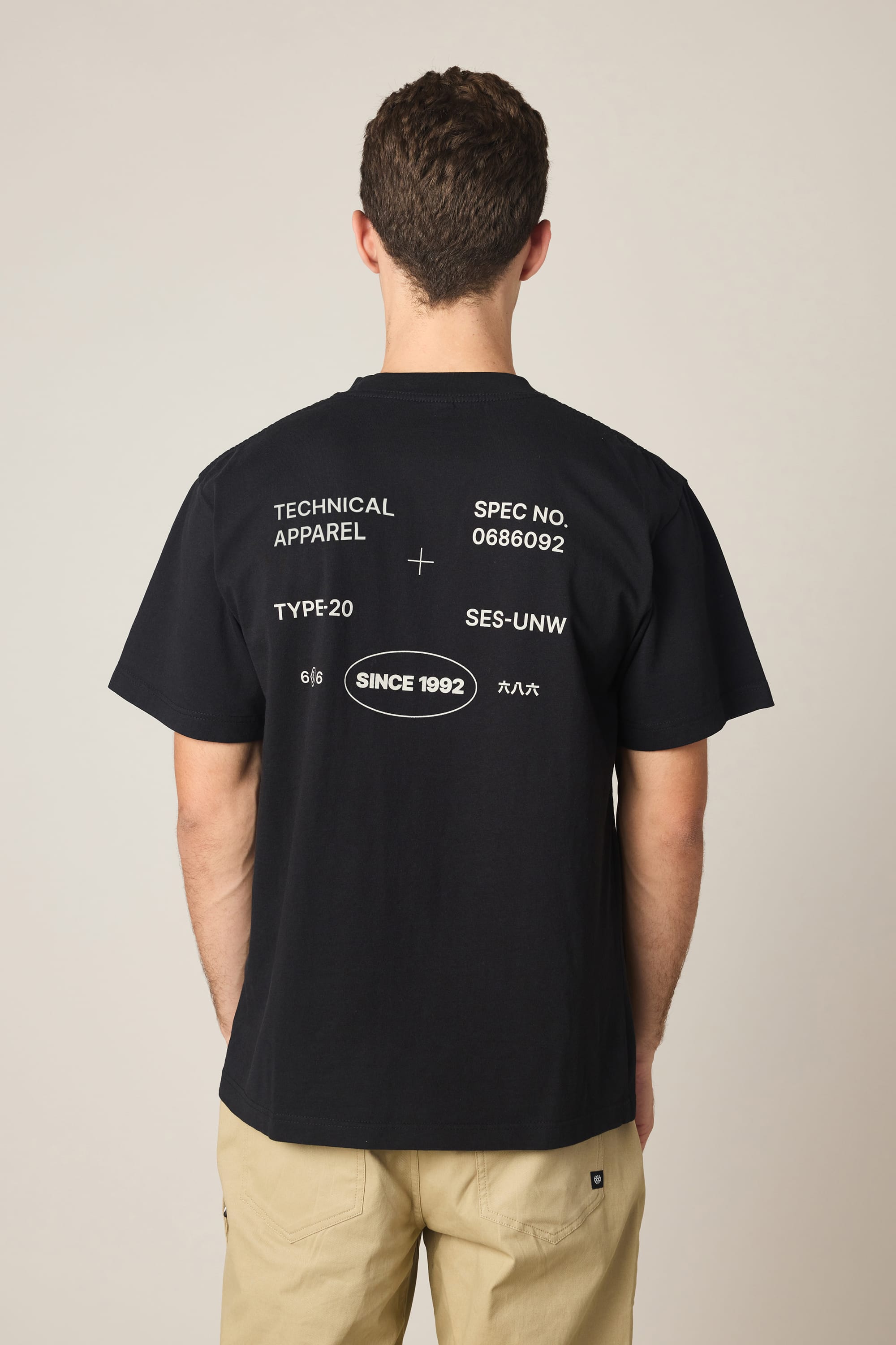 A man with short brown hair wears the 686 Men's Unwork Spec Relaxed SS Tee—a black cotton shirt with "TECHNICAL APPAREL" and "SINCE 1992" in white graphic text—paired with beige pants, facing away from the camera against a plain background.