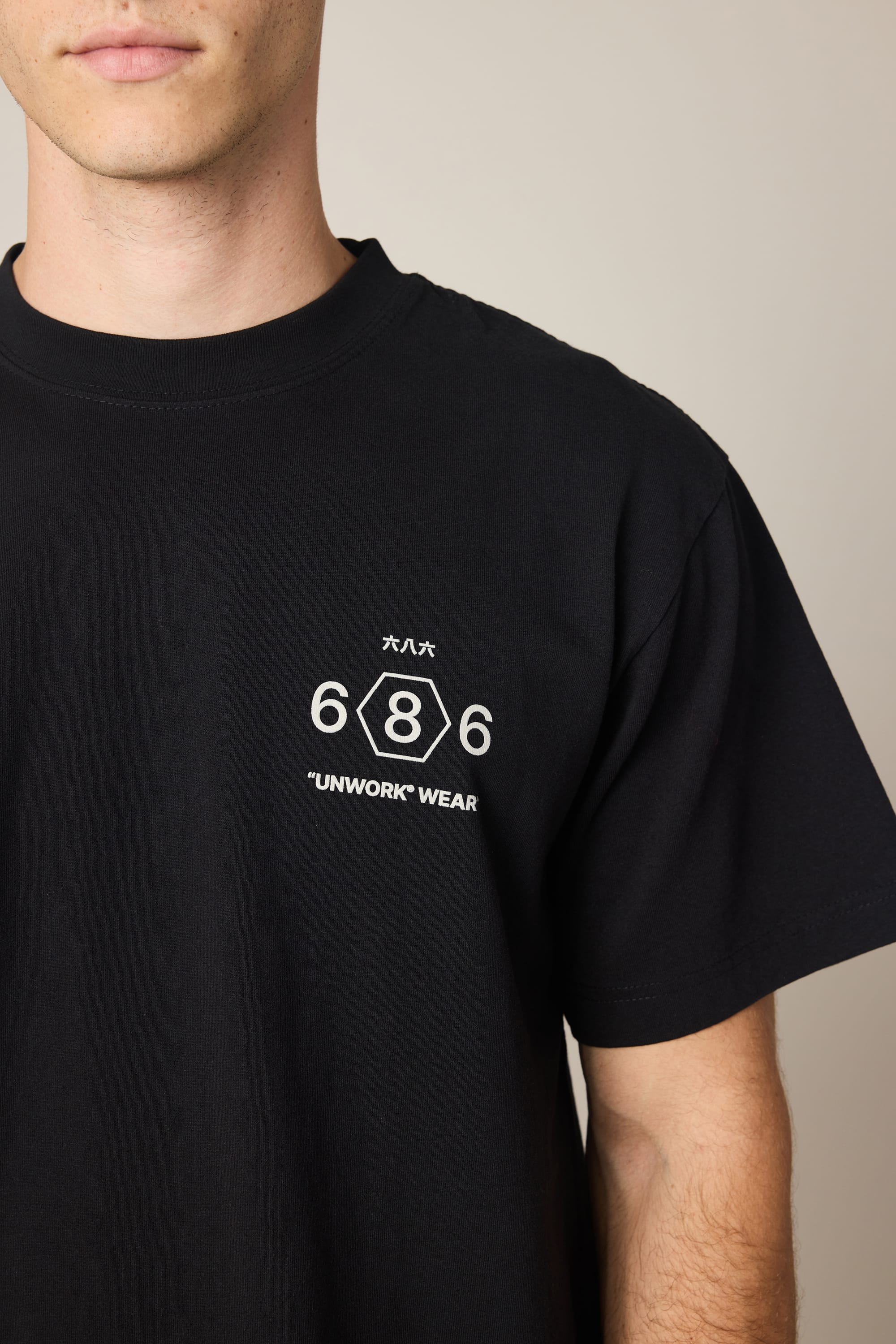 A man wears the 686 Men's Unwork Spec Relaxed SS Tee by 686, a black cotton t-shirt with white prints: "686," a hexagon with an 8, Japanese text, and "UNWORK WEAR." The image shows his upper torso and part of his face.