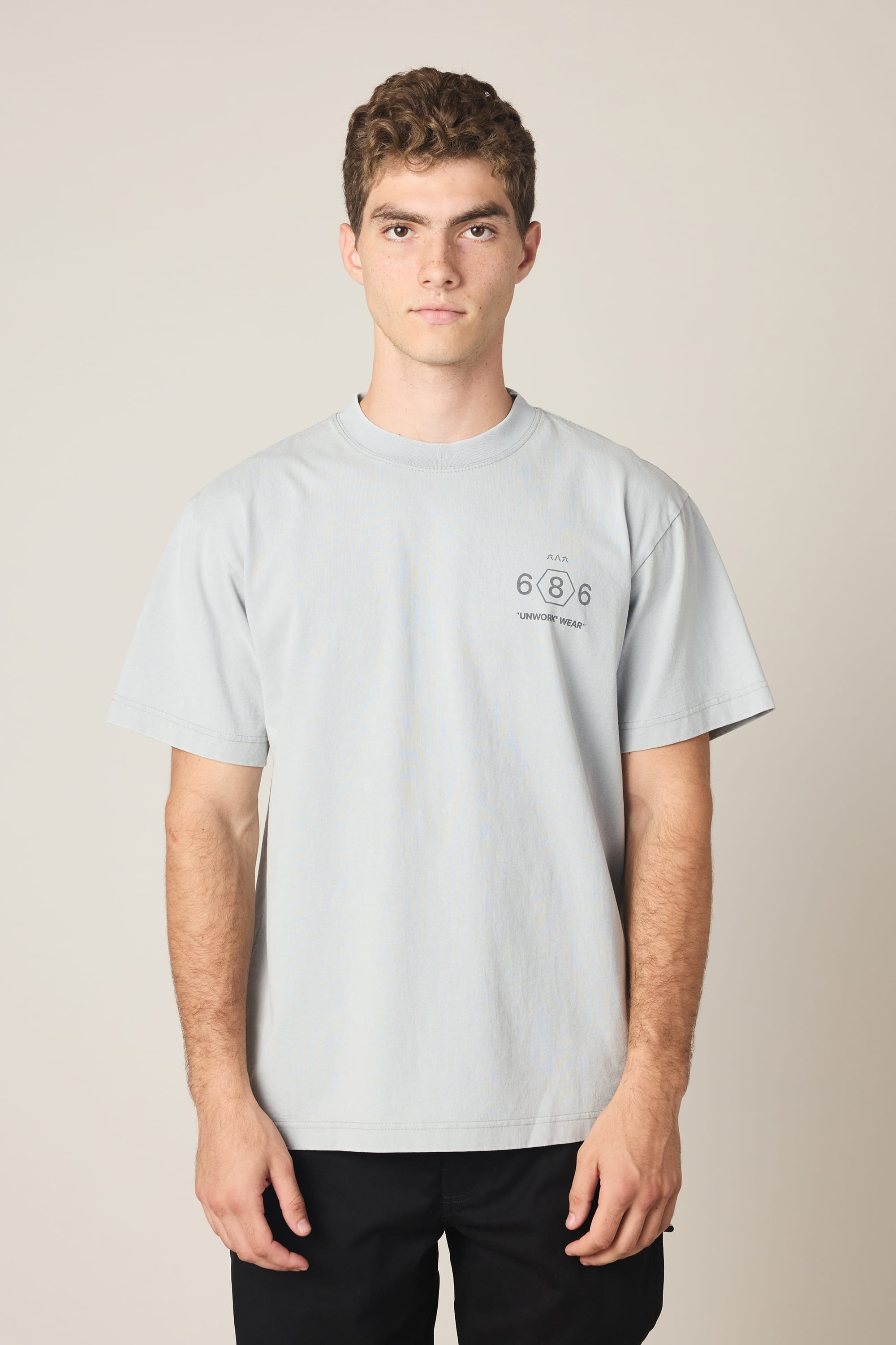 A young man with short curly hair wears the 686 Men's Unwork Spec Relaxed SS Tee in light gray and black pants, standing against a plain background. The 100% cotton tee by 686 features small printed text and numbers on the left chest.