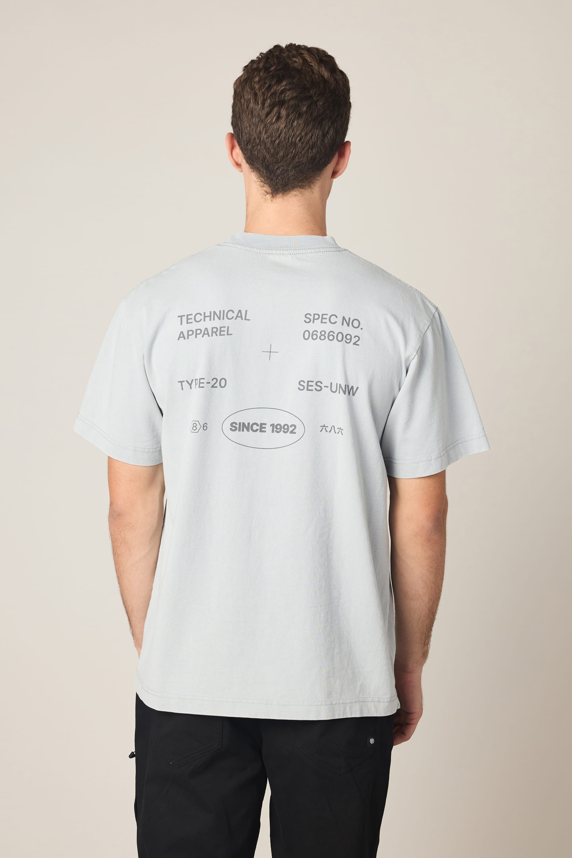A man stands facing away, wearing the 686 Men's Unwork Spec Relaxed SS Tee by 686, a light gray 100% cotton shirt with black printed text and symbols on the back like “TECHNICAL APPAREL,” “SINCE 1992,” codes, and graphics. He pairs it with black pants.