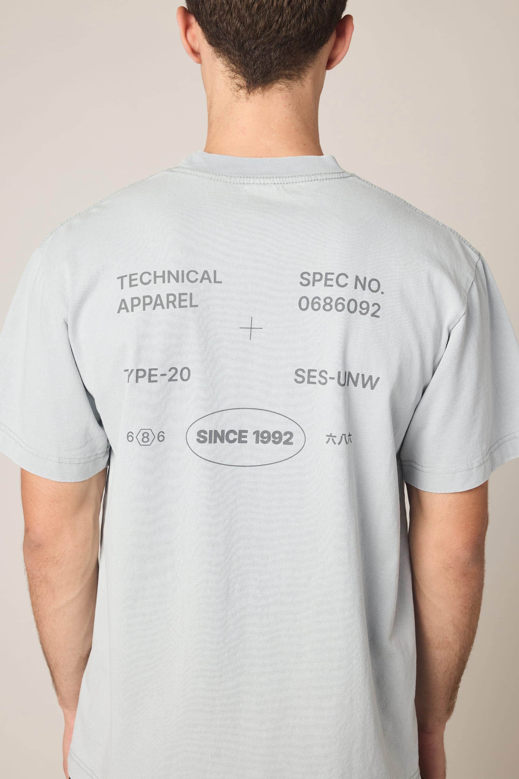 A person is wearing the 686 Men's Unwork Spec Relaxed SS Tee by 686, a light gray t-shirt made from 100% cotton, featuring black technical-style text on the back with phrases like TECHNICAL APPAREL, SINCE 1992, and various codes and symbols.
