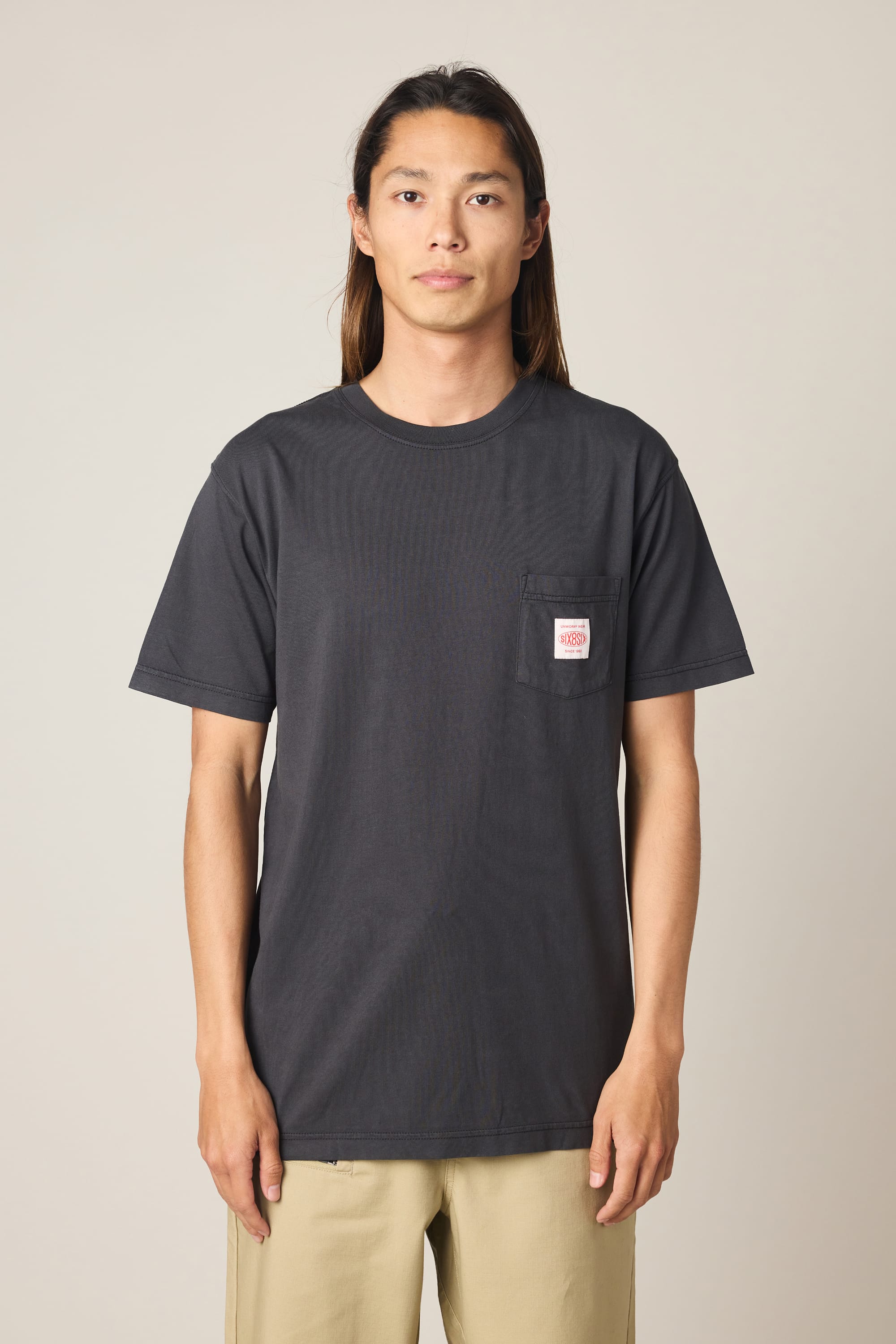 A person with long brown hair wears the 686 Men's Unwork Authentic SS Pocket Tee by 686, a black 100% cotton men's t-shirt featuring a small white and red logo on the chest pocket, paired with beige pants. They stand against a plain light background, facing the camera.