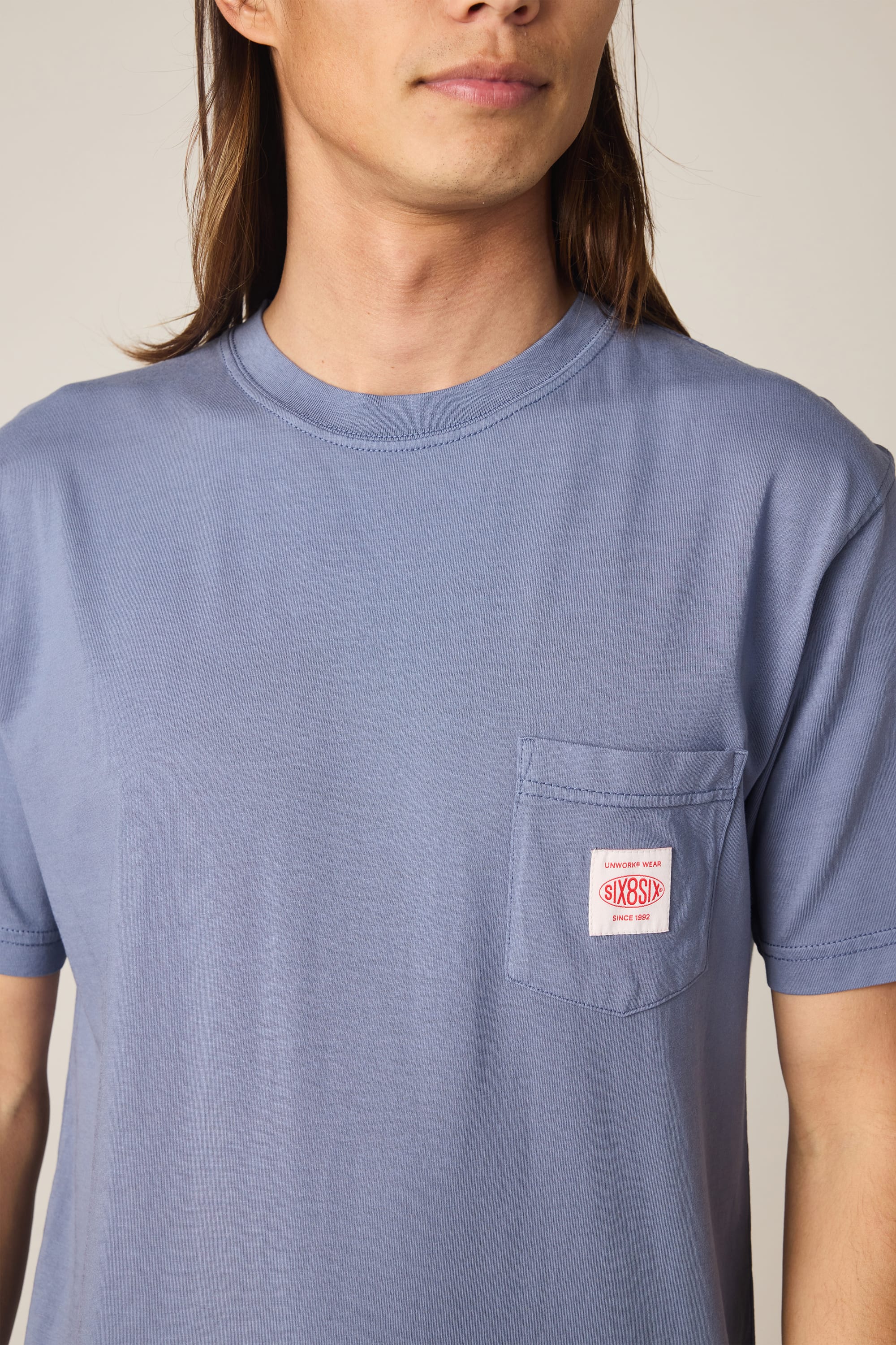 A person wearing the 686 Men's Unwork Authentic SS Pocket Tee—a light blue, 100% cotton men’s t-shirt with a relaxed fit—features a chest pocket adorned with a small red and white SIXSIX patch. The person's face is partially out of frame.