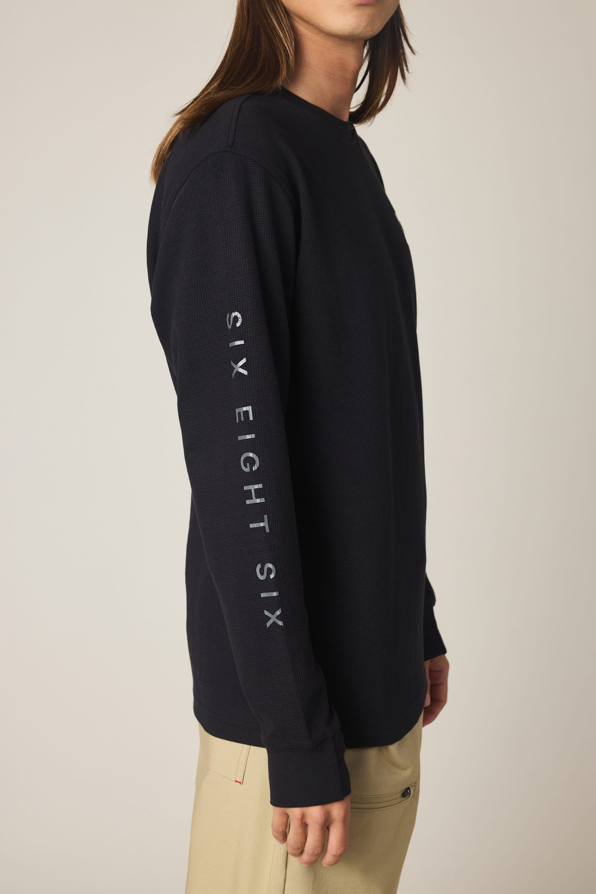 A man wears the 686 Men's Let's Chill Waffle LS Crew, a black polyester long-sleeve t-shirt with “SIX EIGHT SIX” printed in white vertically on the left sleeve, paired with beige pants against a plain background.