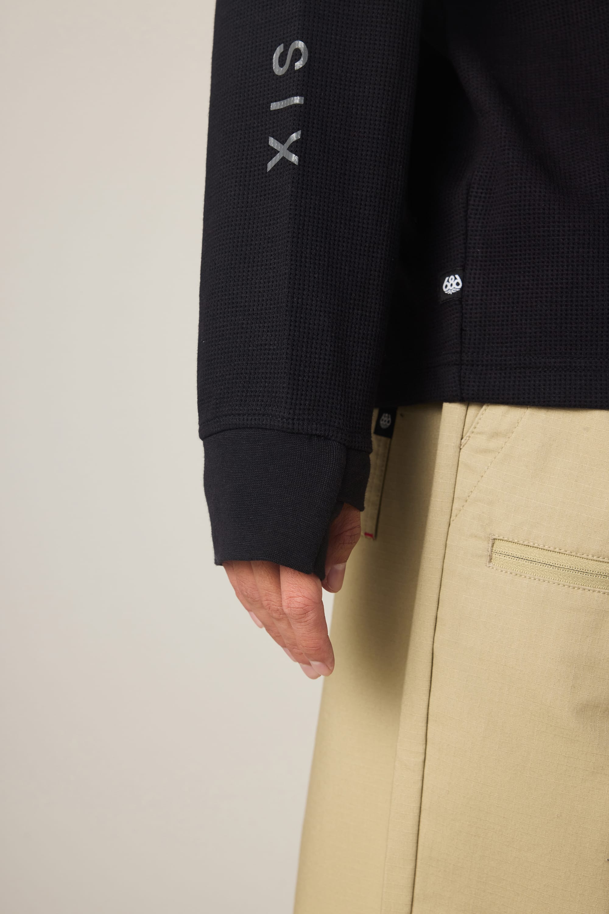 A person wears the 686 Men's Let's Chill Waffle LS Crew by 686, a black long sleeve polyester t-shirt with text on the sleeve and beige pants, shown waist down with their hand relaxed at their side against a plain background.