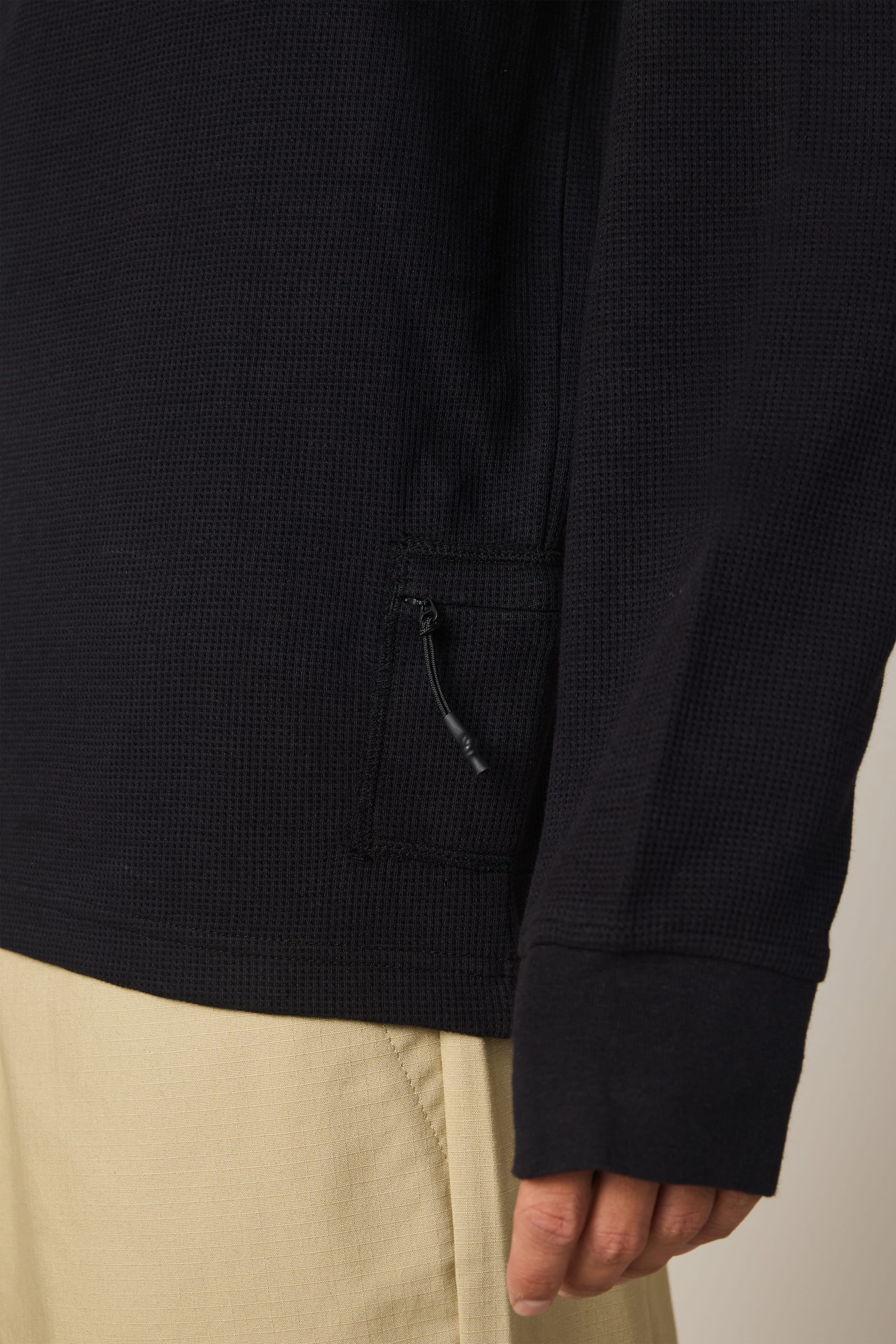 A close-up of a person wearing the 686 Men's Let's Chill Waffle LS Crew in black, featuring a textured design and small zippered pocket near the hem, paired with light beige pants. Only the lower torso and hand are visible.