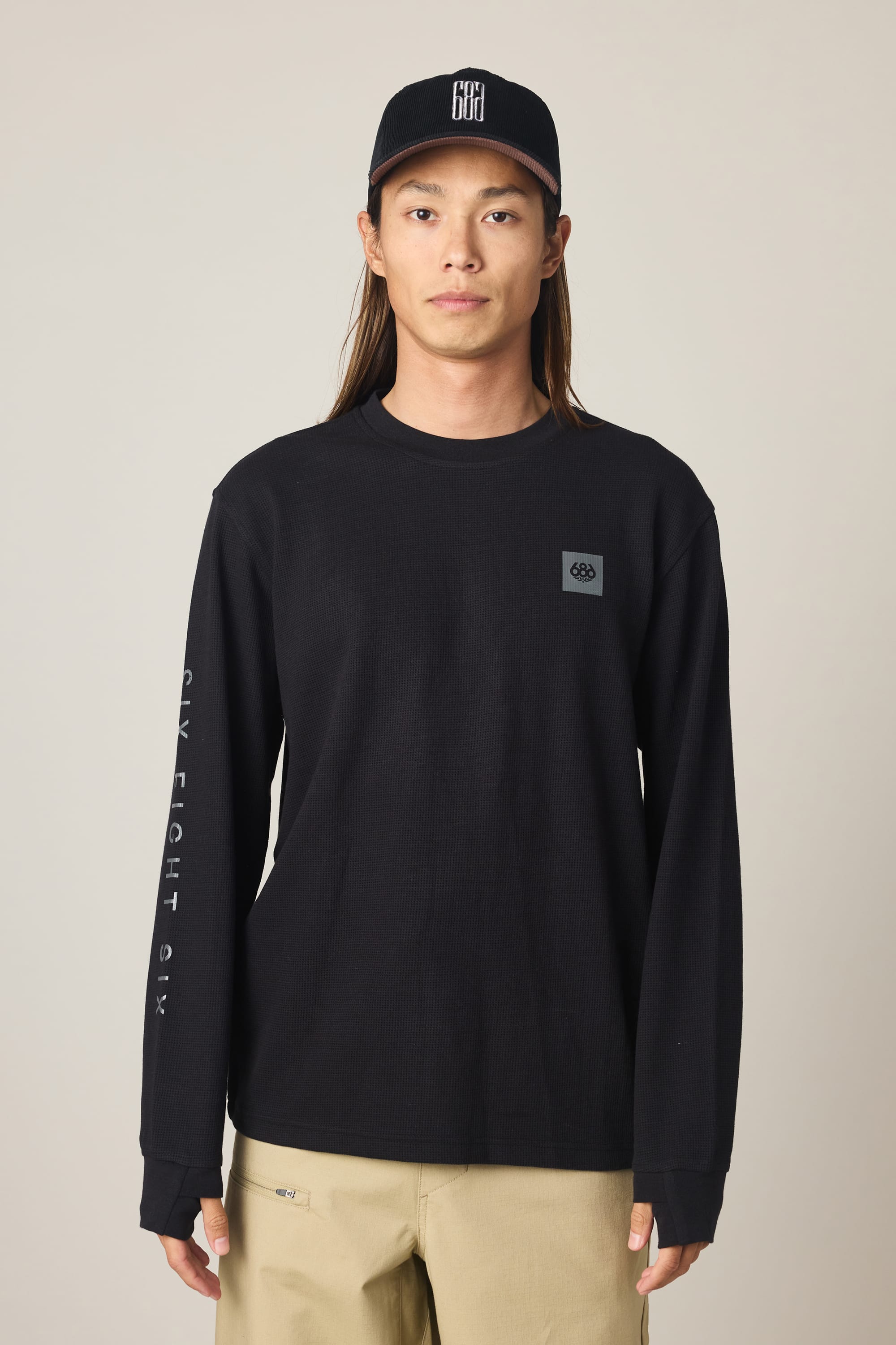 A person in the 686 Men's Let's Chill Waffle LS Crew by 686, featuring a small chest logo and text on the sleeve, paired with beige pants and a black baseball cap, stands facing forward against a plain light background.