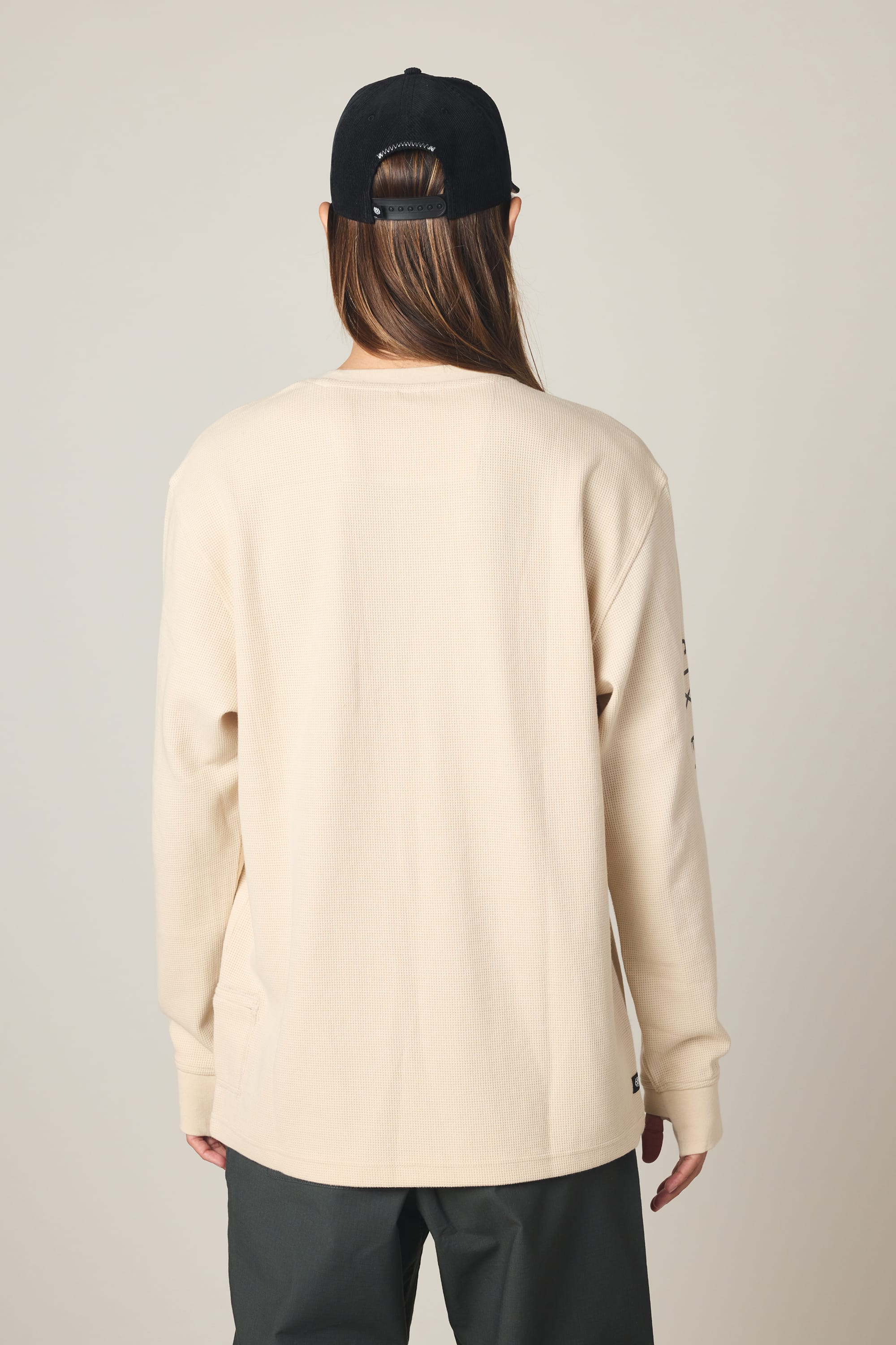 A person faces away from the camera, wearing the 686 Men's Let's Chill Waffle LS Crew in cream, dark pants, and a black baseball cap with long brown hair visible through the back against a plain light background.