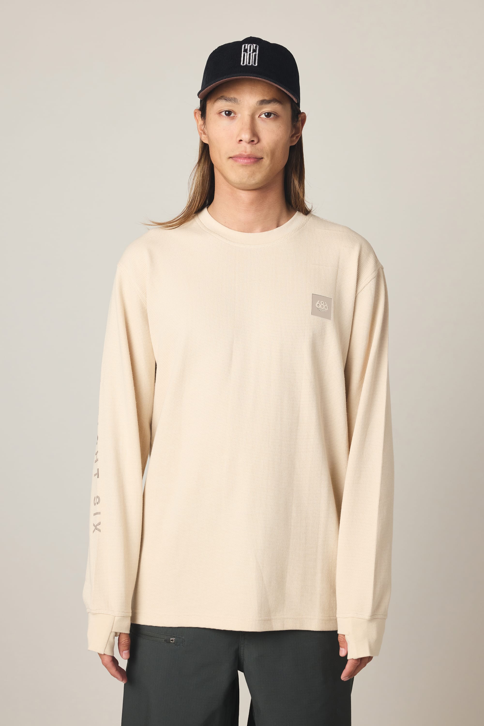 A person with long hair is wearing a black baseball cap, the 686 Men's Let's Chill Waffle LS Crew by 686 in beige, and dark pants, standing against a plain light background.
