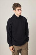 A young man with short curly hair wears a black 686 Men's Let's Chill Waffle Hoody, paired with khaki pants, standing against a plain light background and looking slightly to the side with a neutral expression.