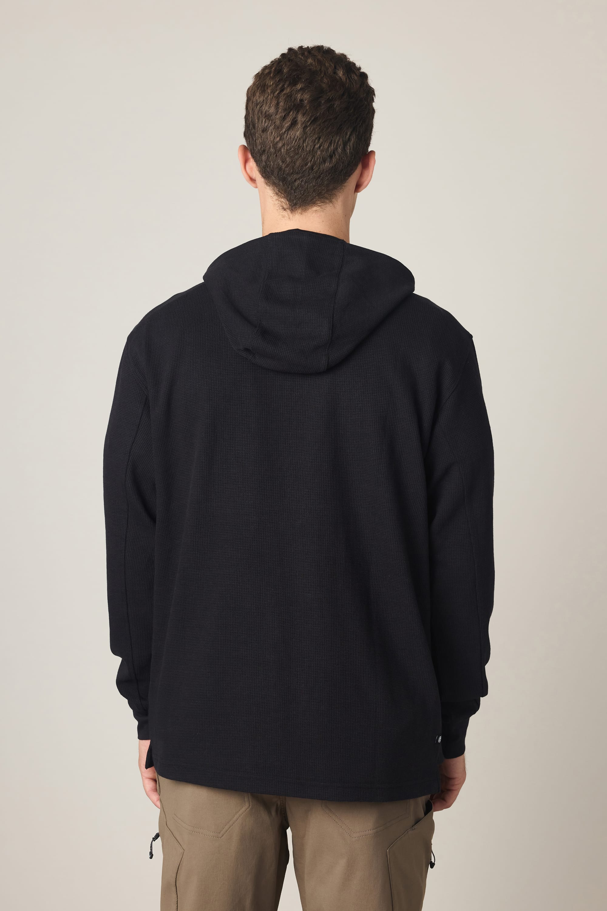 A person with short curly hair stands with their back to the camera, wearing the 686 Men's Let's Chill Waffle Hoody by 686 and light brown pants against a plain light background.