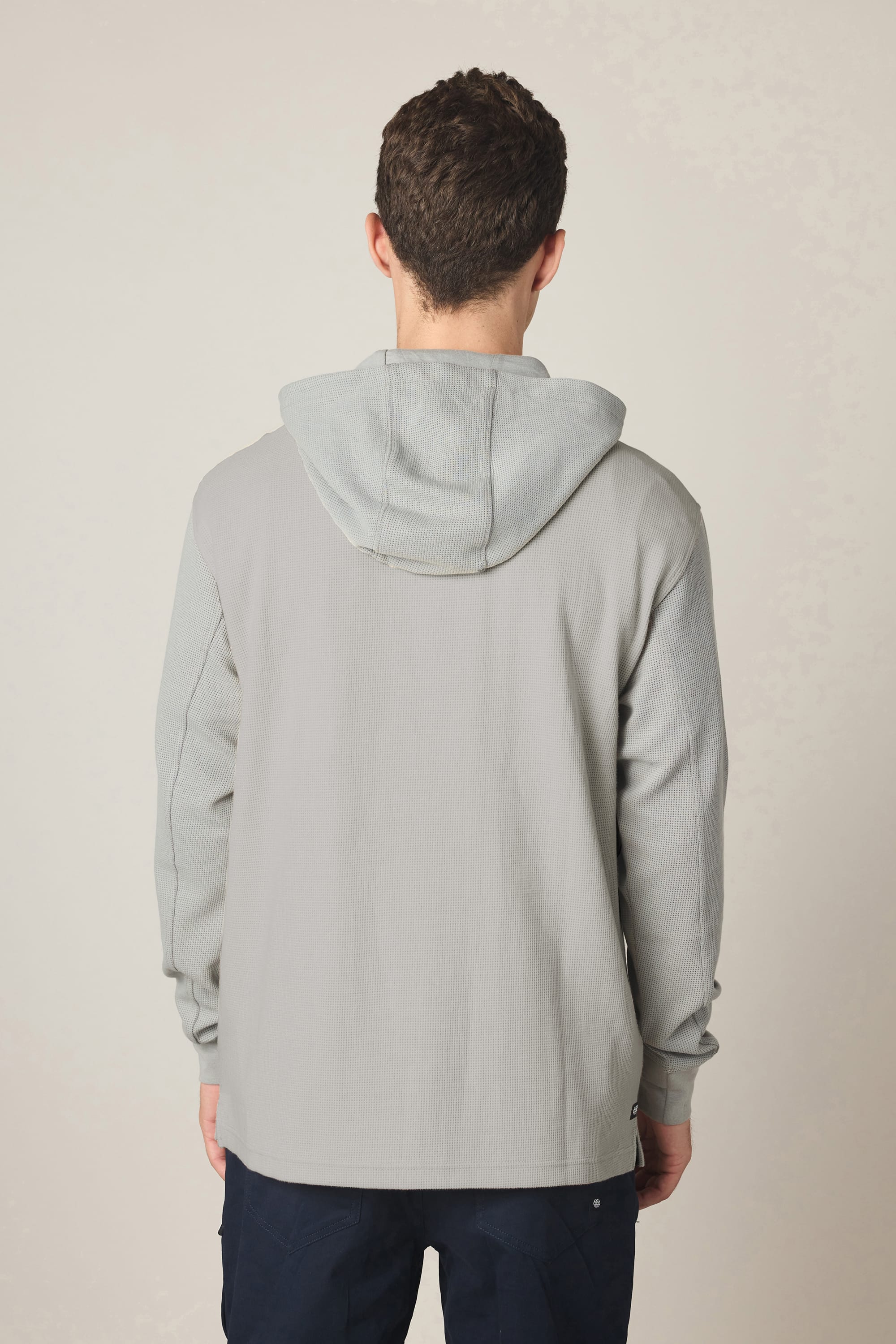 A person with short brown hair stands with their back to the camera, wearing a light gray 686 Men's Let's Chill Waffle Hoody by 686 and dark blue pants against a plain, light background.