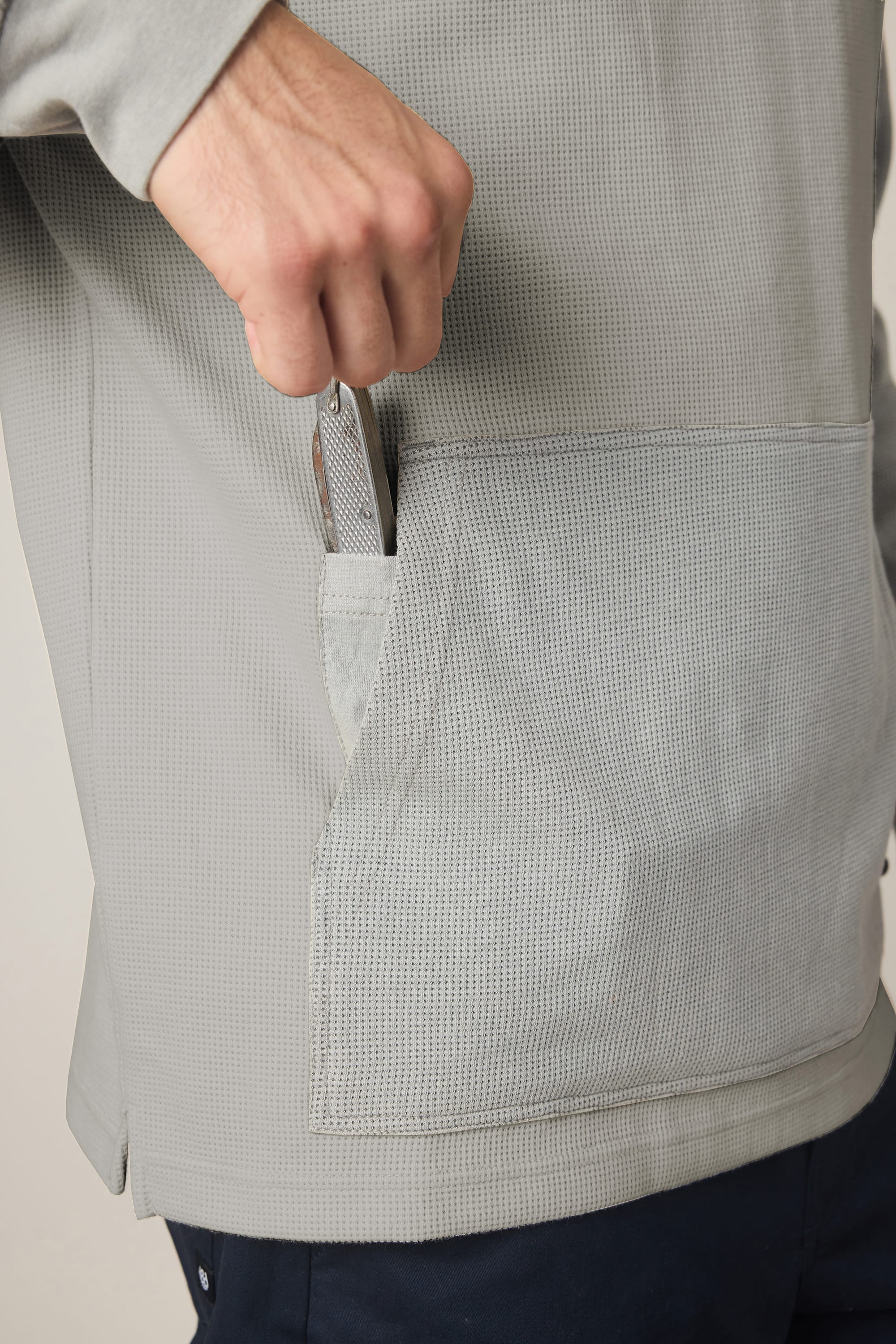 A person wearing a 686 Men's Let's Chill Waffle Hoody in light gray is placing a folded pocket knife into its front pocket. Only the person's hand and torso are visible.