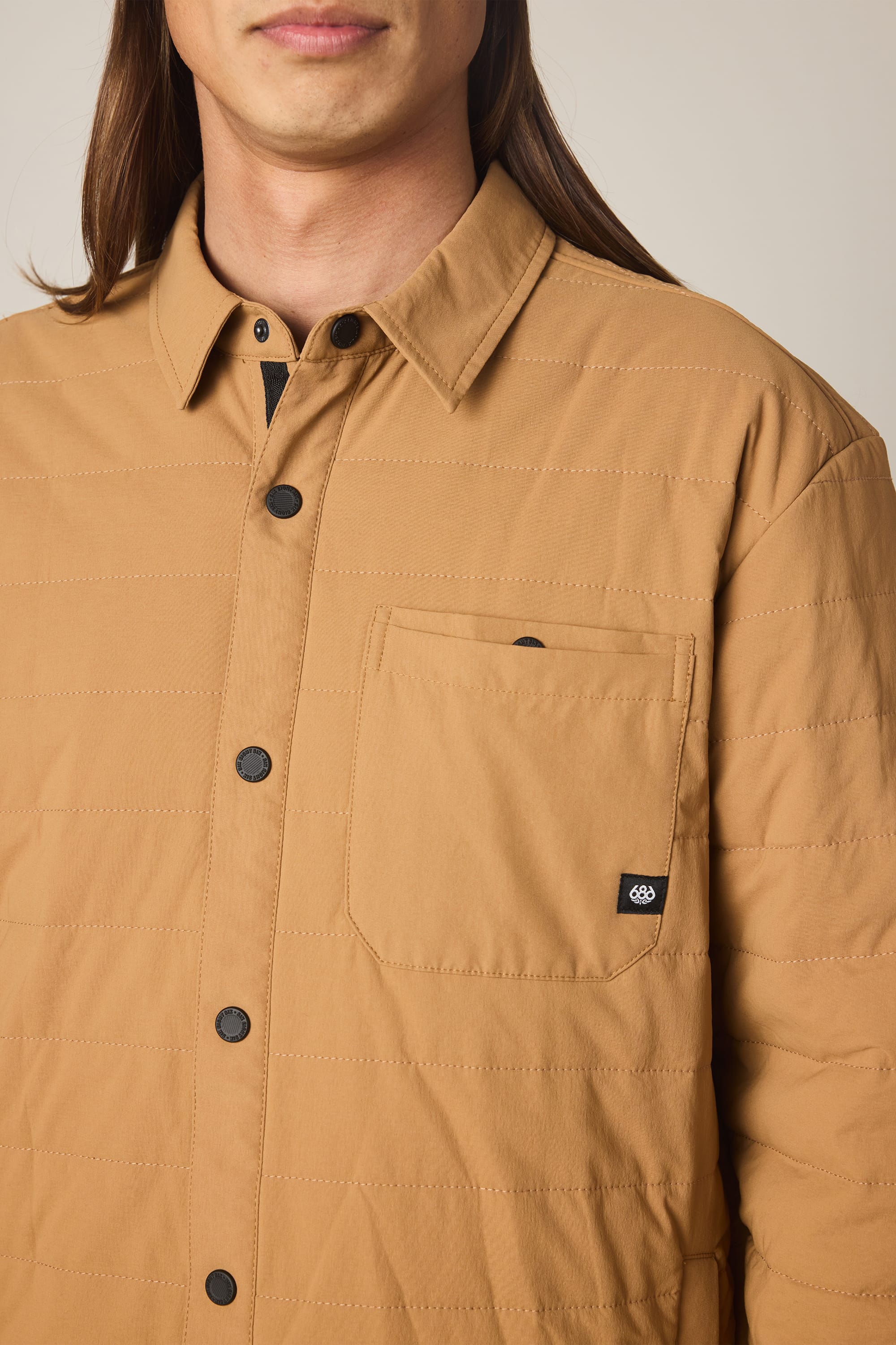 A person wears the 686 Men's Thermaldry Insulated Shacket, a tan quilted jacket with black buttons and a chest pocket featuring a small black tag. The image focuses on the upper torso only.